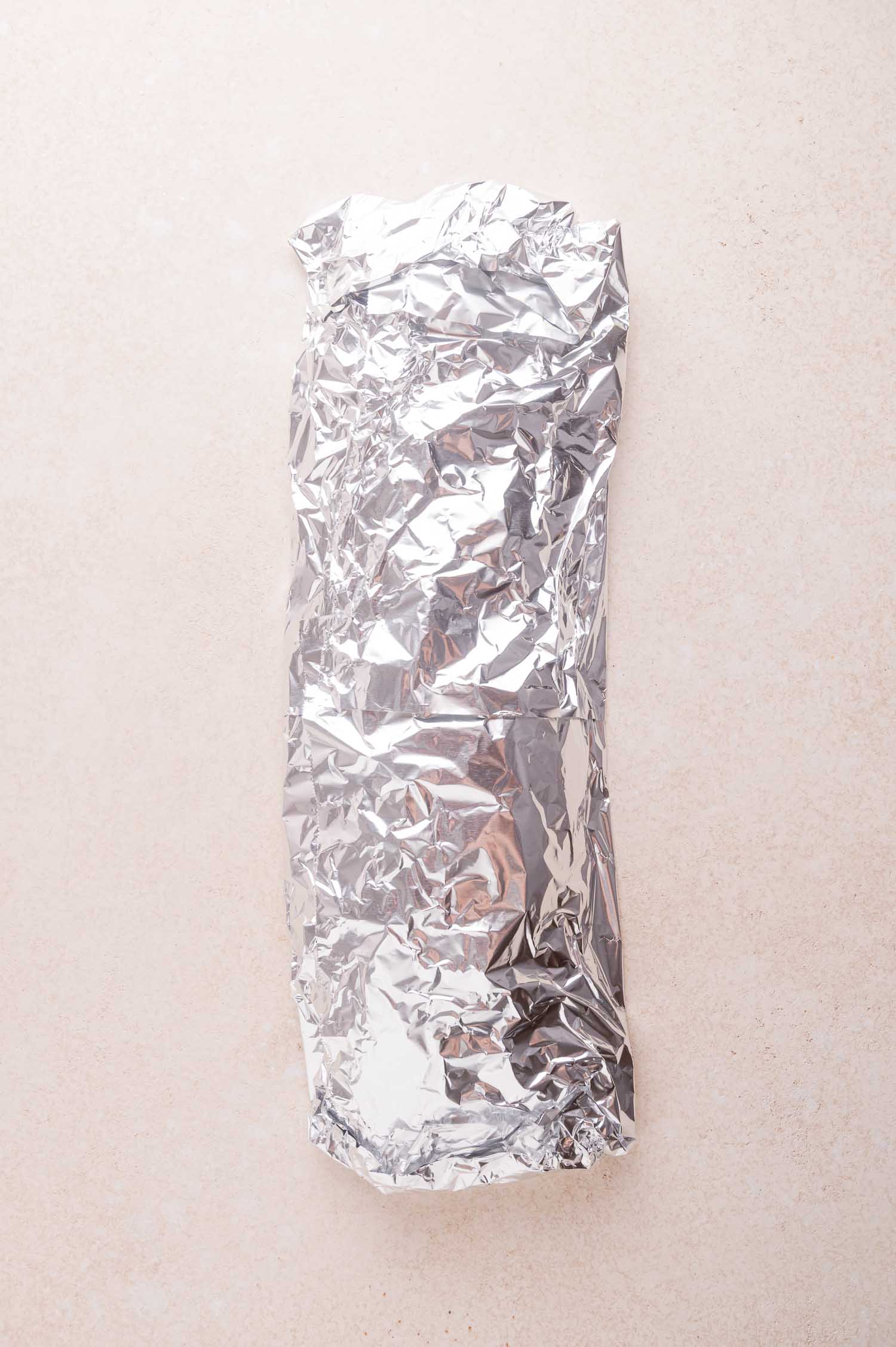 A rack of ribs wrapped tightly in crinkled aluminum foil, placed on a light-colored surface.