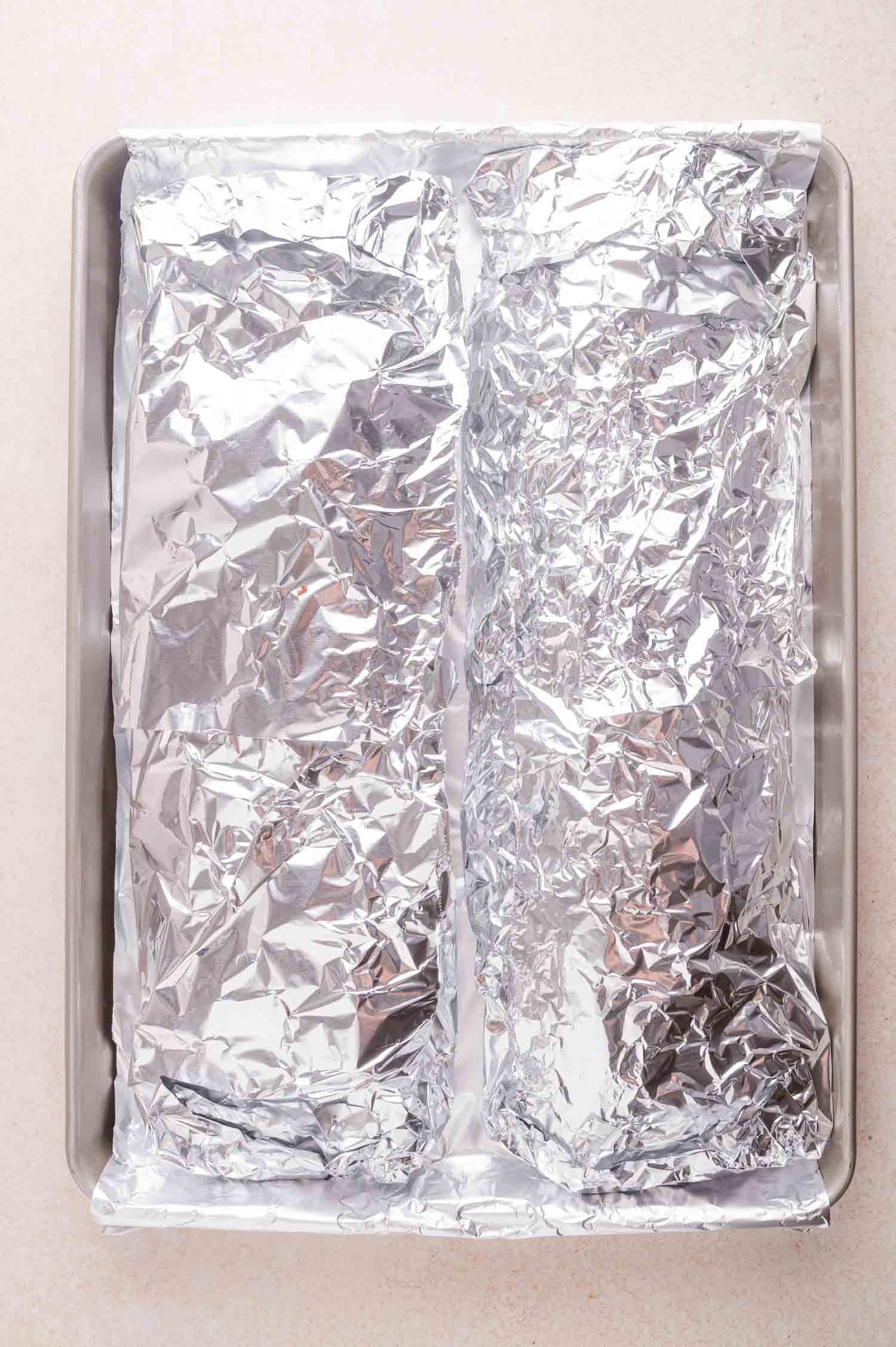 A baking sheet with two racks of ribs wrapped in aluminum foil, placed side by side on a light-colored countertop.