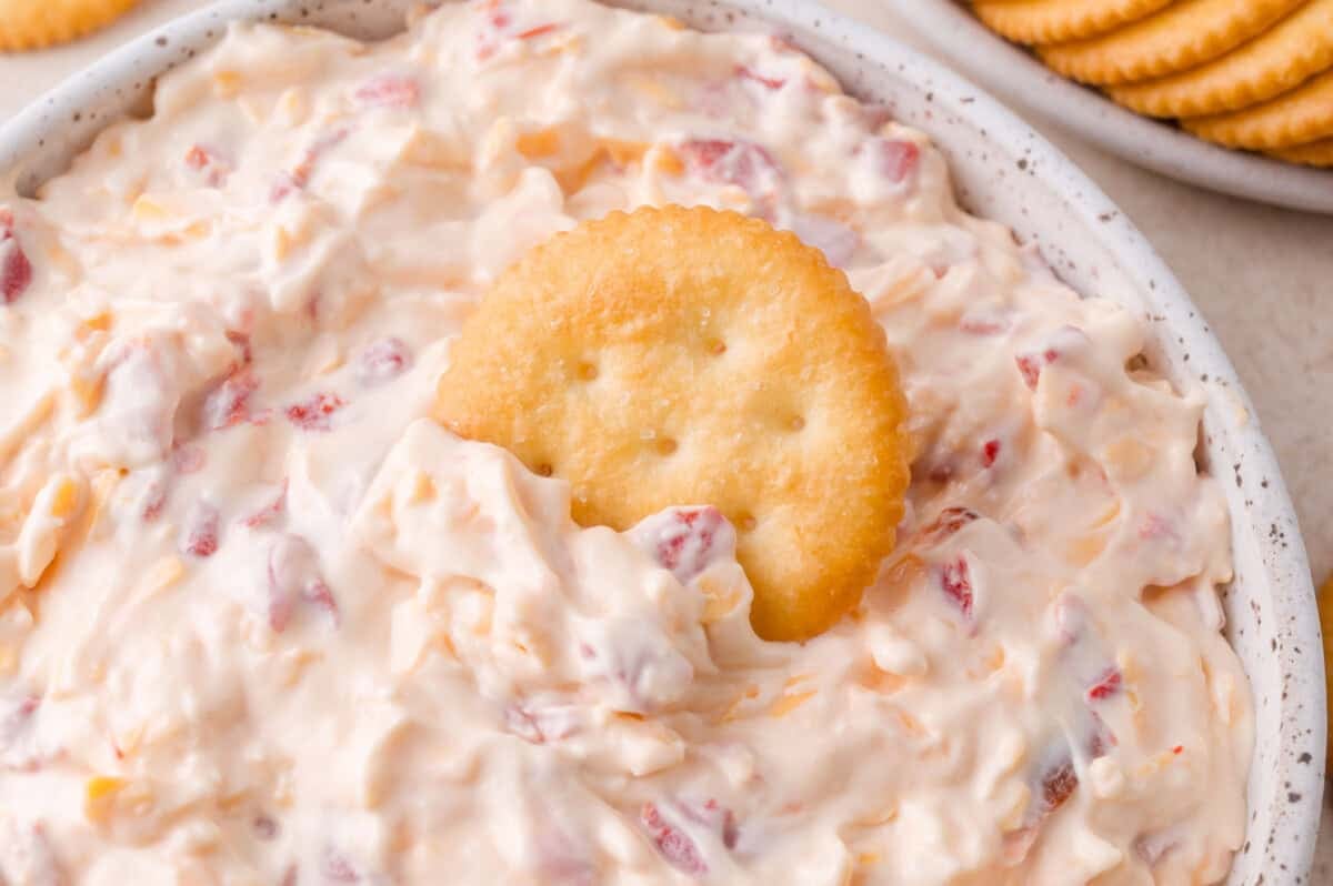 A round cracker is partially dipped in a creamy cheese and pimento spread, with more crackers visible in the background.