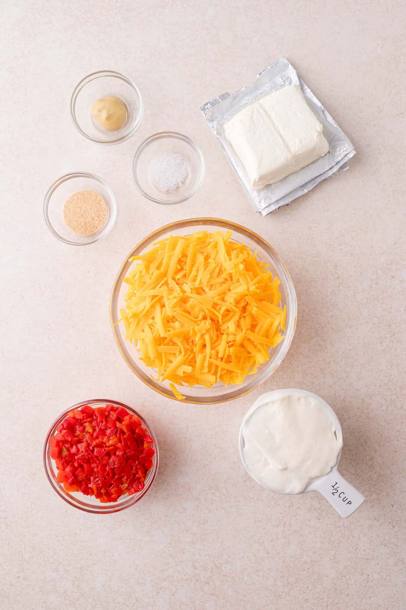 Ingredients for a cheese dip recipe arranged on a countertop, including shredded cheddar, cream cheese, diced pimentos, mayonnaise, mustard, salt, and onion powder.