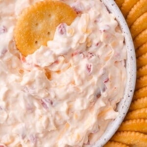 A close-up of a bowl filled with creamy pimento cheese dip containing red pimento pieces, surrounded by round crackers, with one cracker dipped in the mixture.