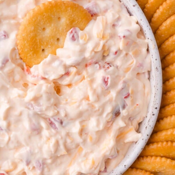 A close-up of a bowl filled with creamy pimento cheese dip containing red pimento pieces, surrounded by round crackers, with one cracker dipped in the mixture.