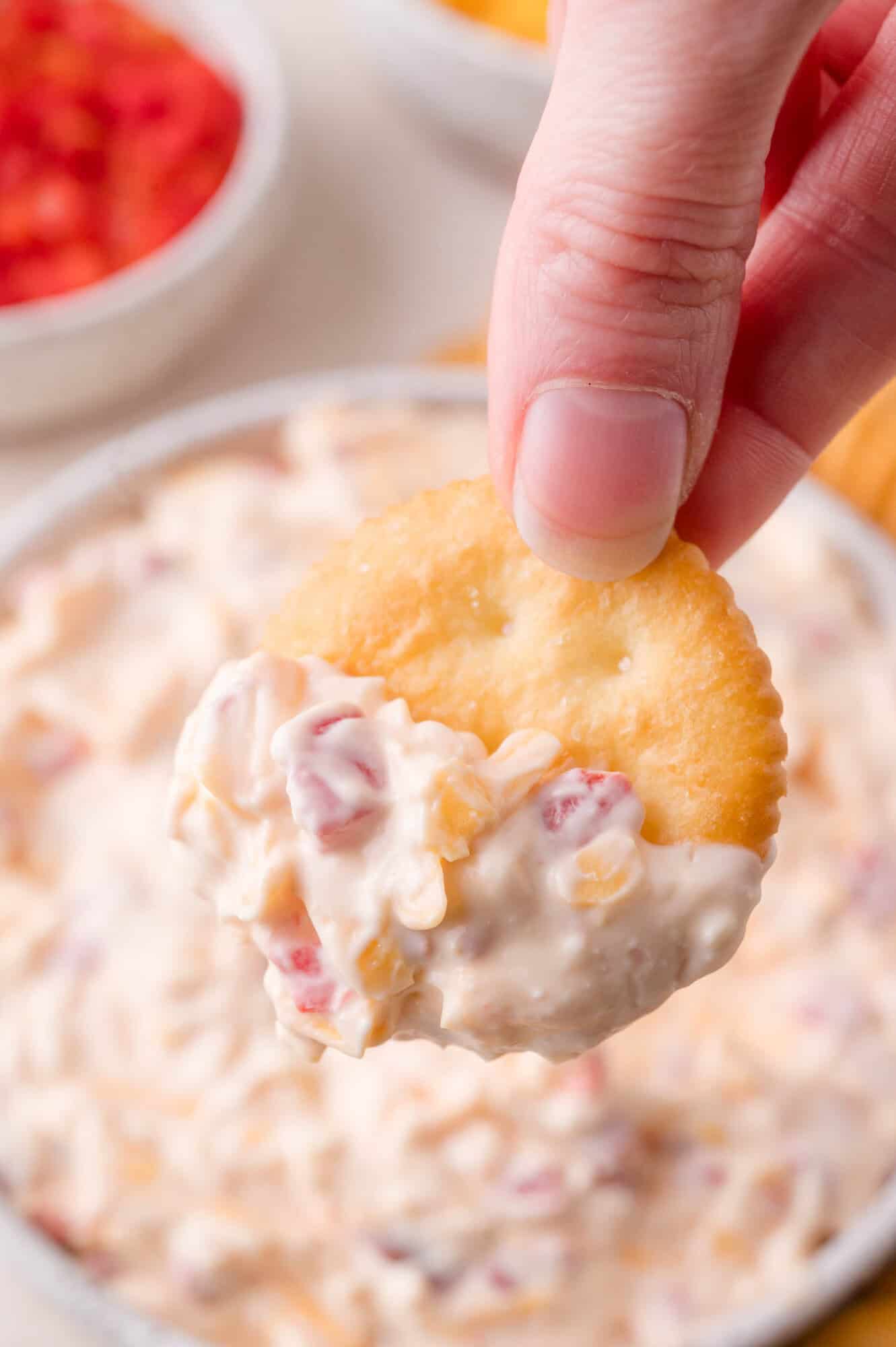 A hand holds a round cracker dipped in a creamy pimento cheese with visible bits of red and yellow ingredients.