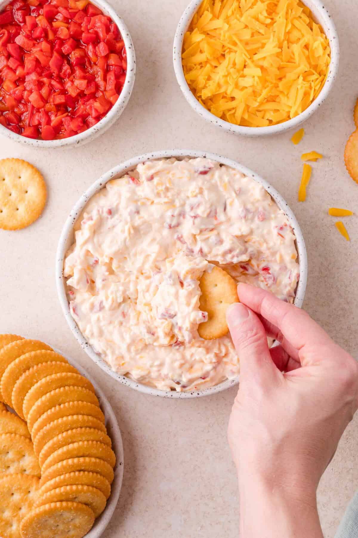 A hand dips a round cracker into a bowl of creamy pimento cheese, surrounded by bowls of shredded cheese, diced pimentos, and more crackers.