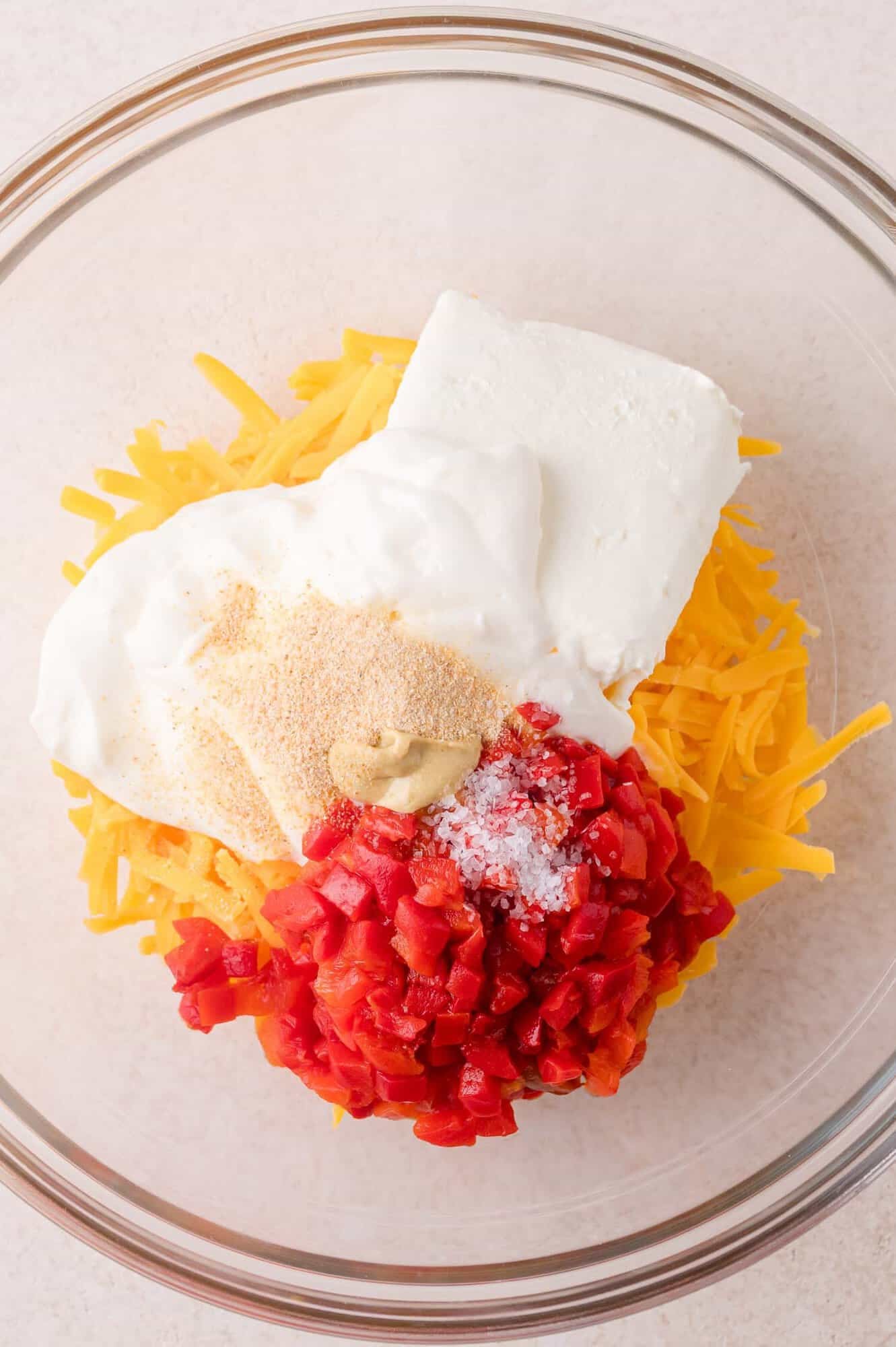 A glass bowl containing shredded cheddar cheese, cream cheese, diced pimentos, mayonnaise, mustard, garlic powder, and salt.