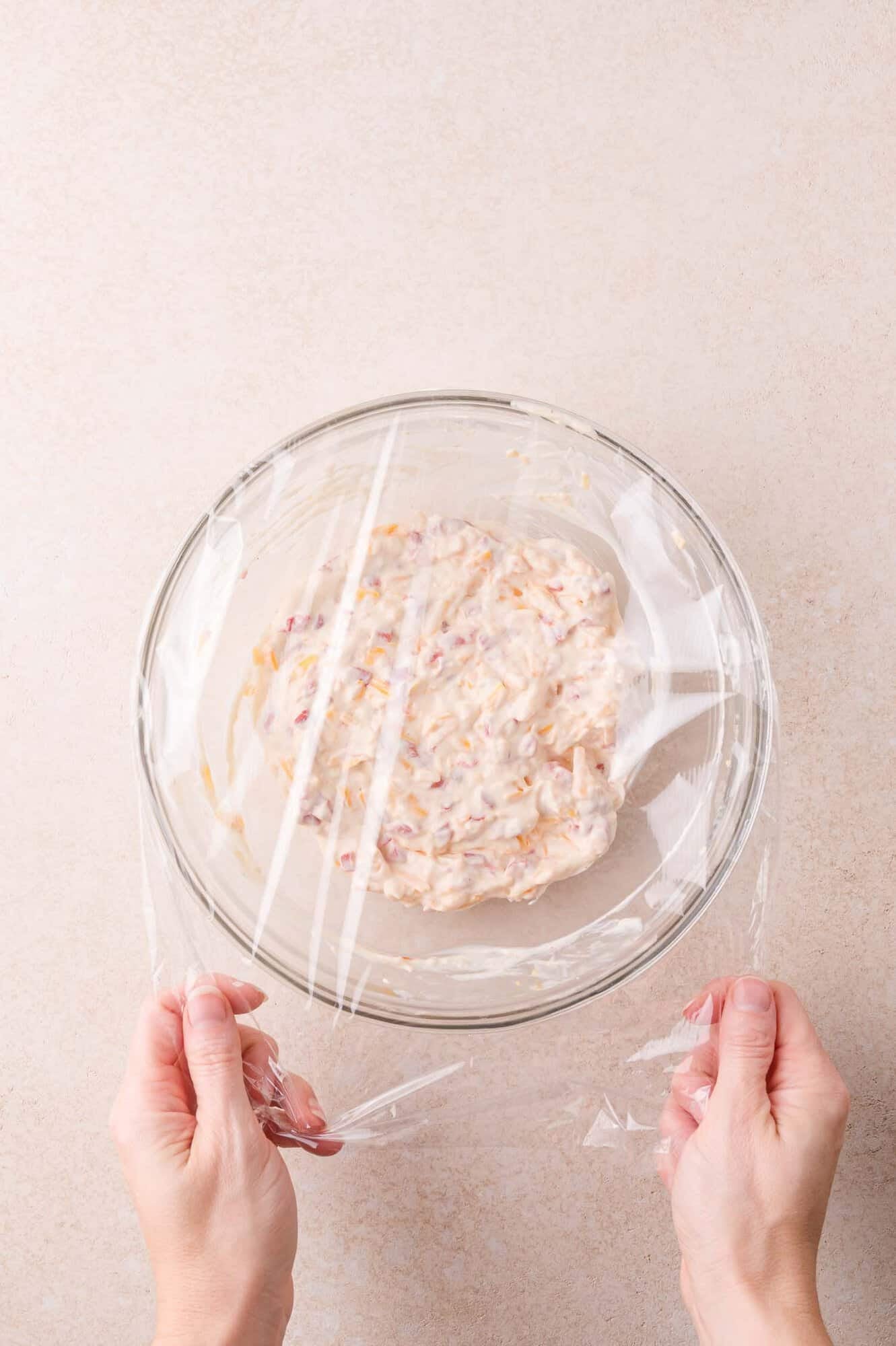 Hands covering a glass bowl filled with a pimento cheese with plastic wrap.