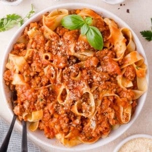 A bowl of pappardelle pasta topped with turkey bolognese sauce, garnished with grated cheese and fresh basil, with a fork and spoon resting inside.