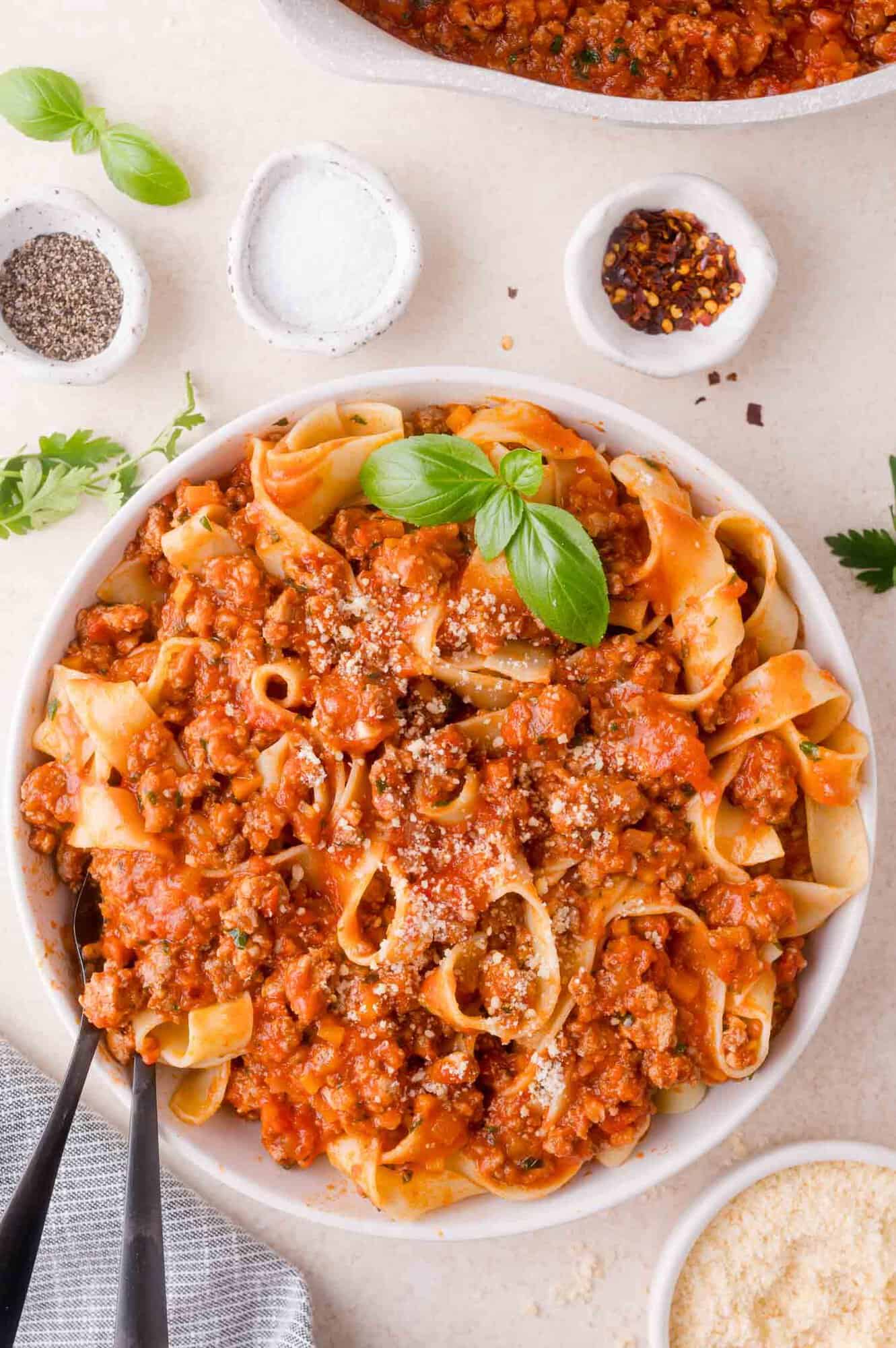 A bowl of pappardelle pasta topped with bolognese sauce, grated cheese, and a basil garnish, with bowls of seasonings and grated cheese on the side.
