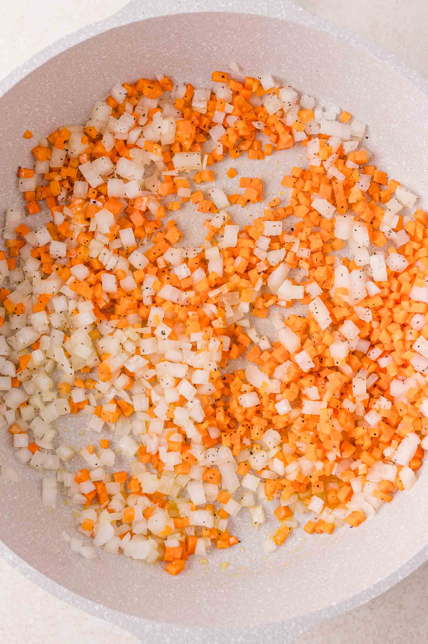 Diced carrots and onions being sautéed in a pan with visible black pepper seasoning.