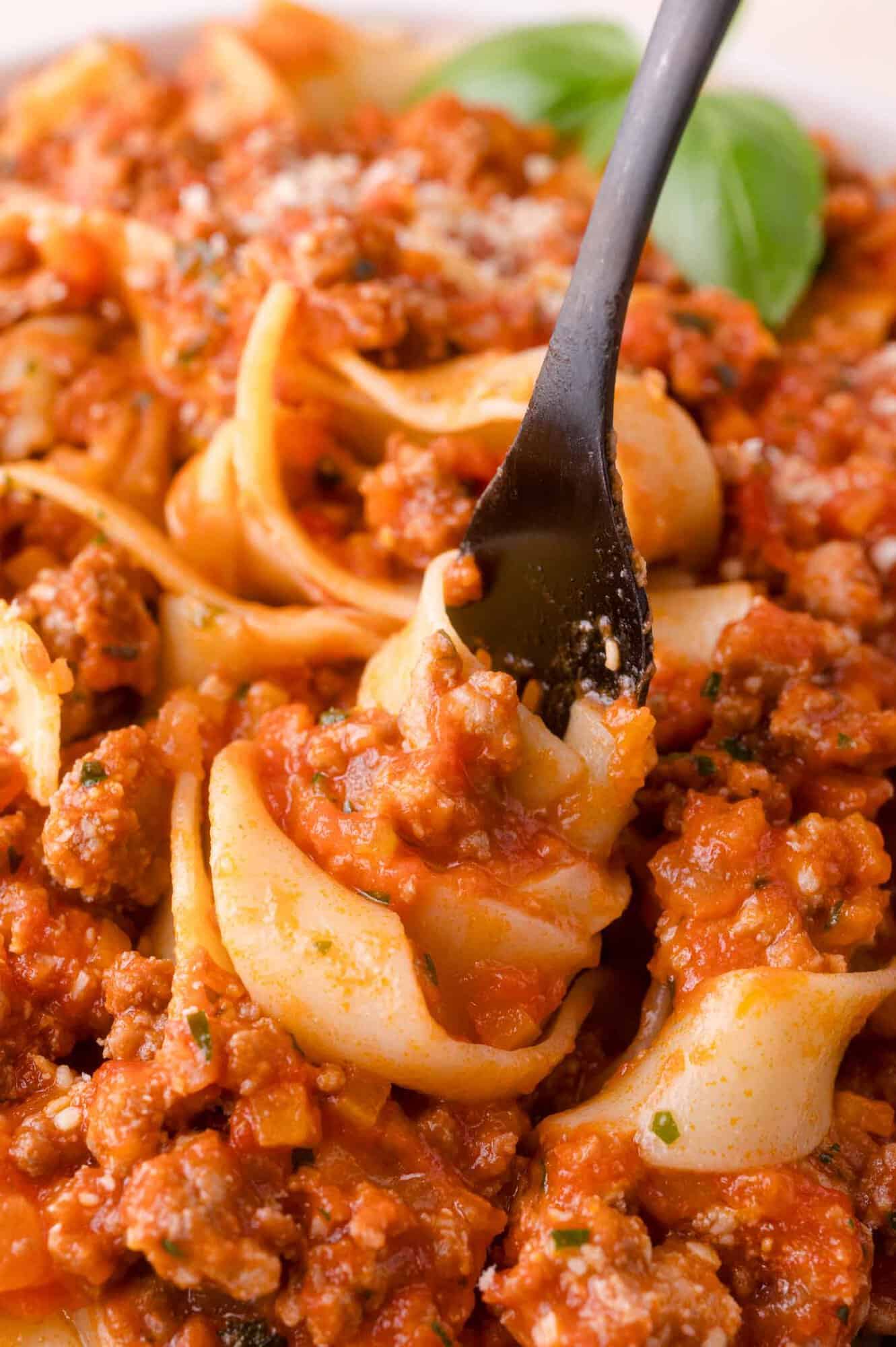 A close-up of a fork twirling pasta with a thick tomato and ground meat sauce, garnished with grated cheese and herbs.