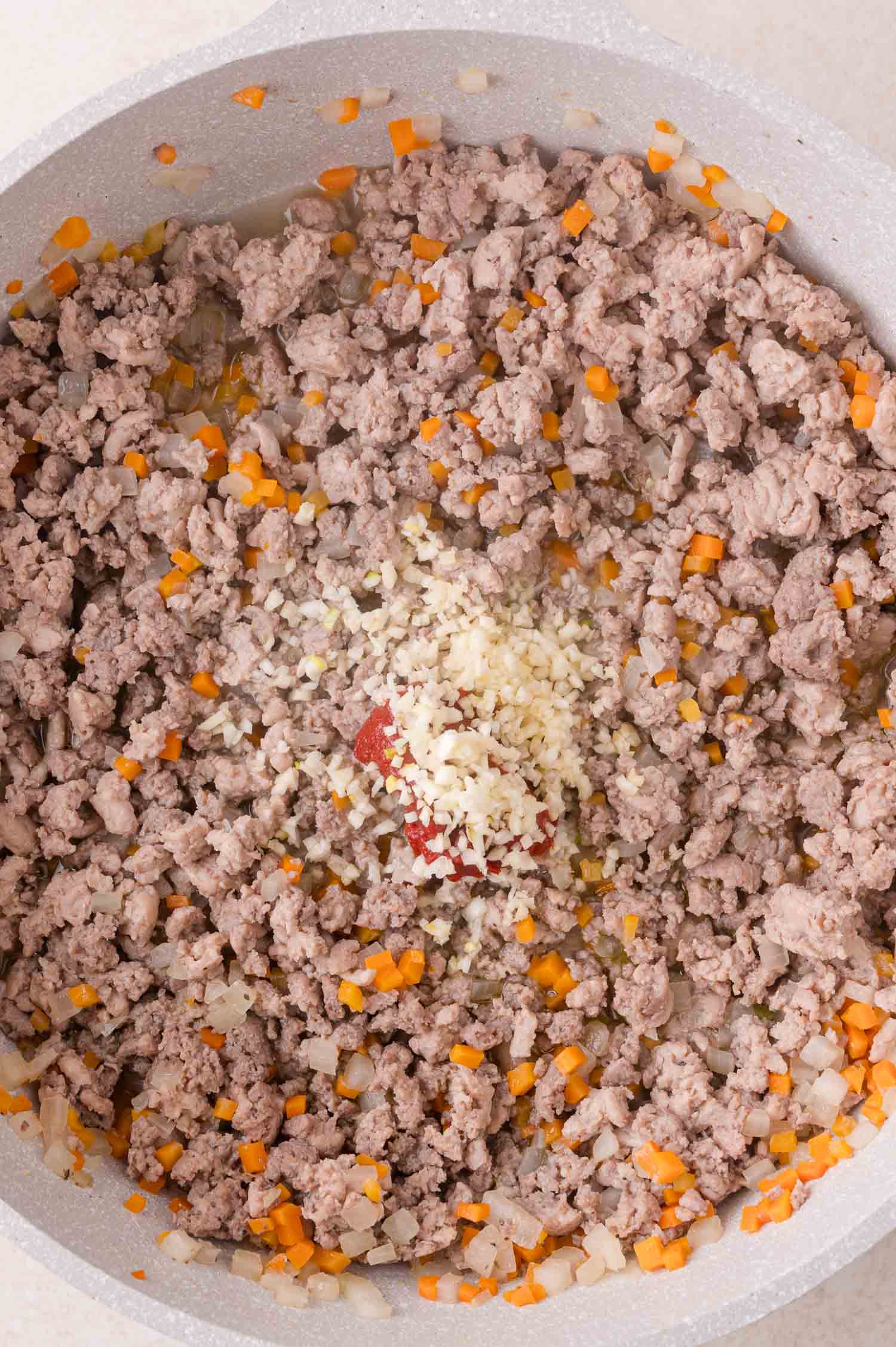 Ground meat, diced carrots, onions, and a dollop of tomato paste topped with minced garlic cooking in a pan.