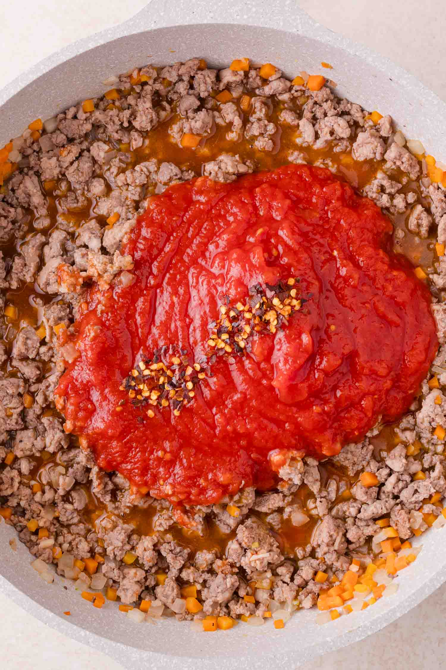 Ground turkey, diced onions, carrots, and tomato sauce topped with red pepper flakes cooking in a skillet.