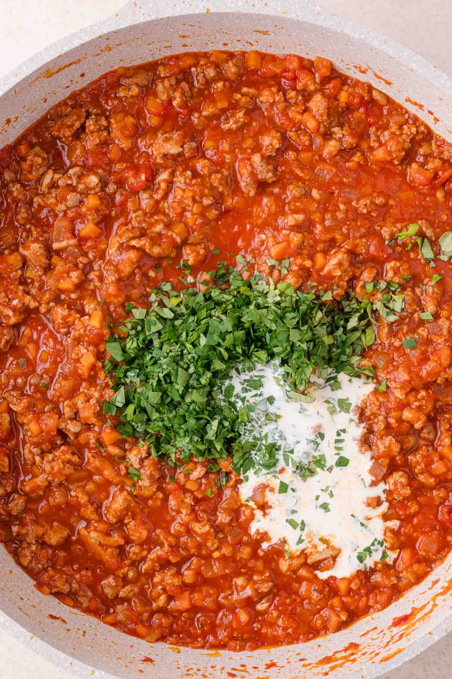 Pan of tomato-based meat sauce with a dollop of cream and a pile of chopped fresh herbs in the center, about to be mixed in.