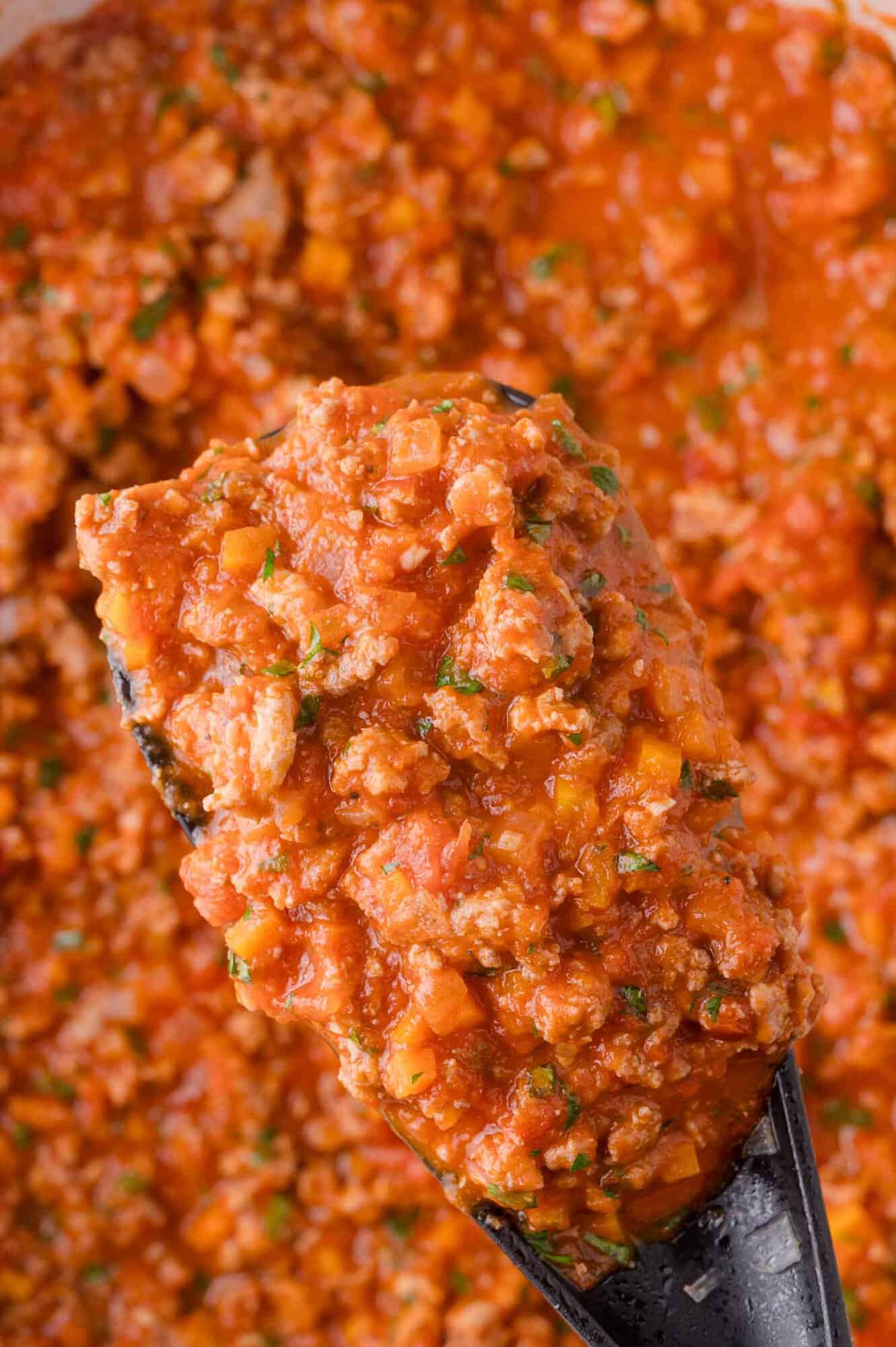 Close-up of a spoonful of turkey bolognese, a chunky tomato-based meat sauce with herbs and vegetables, held above a pot of the same sauce.