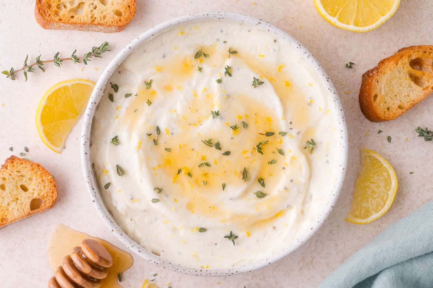 A bowl of whipped ricotta cheese topped with honey, lemon zest, and fresh thyme, surrounded by crostini, lemon slices, and a honey dipper.
