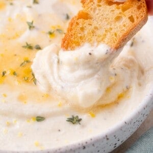 A piece of toasted bread is being dipped into a bowl of creamy whipped ricotta garnished with herbs and lemon zest.