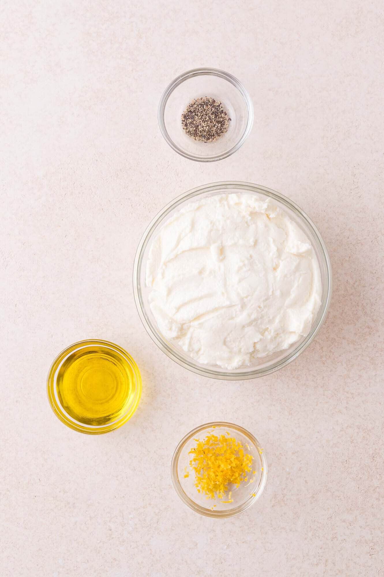 Four small bowls containing black pepper, ricotta cheese, olive oil, and lemon zest are arranged on a light-colored surface.