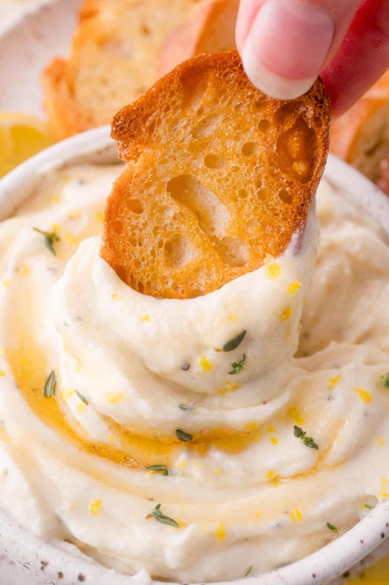 A hand dips a toasted bread slice into a bowl of creamy whipped ricotta, garnished with herbs and a drizzle of oil.