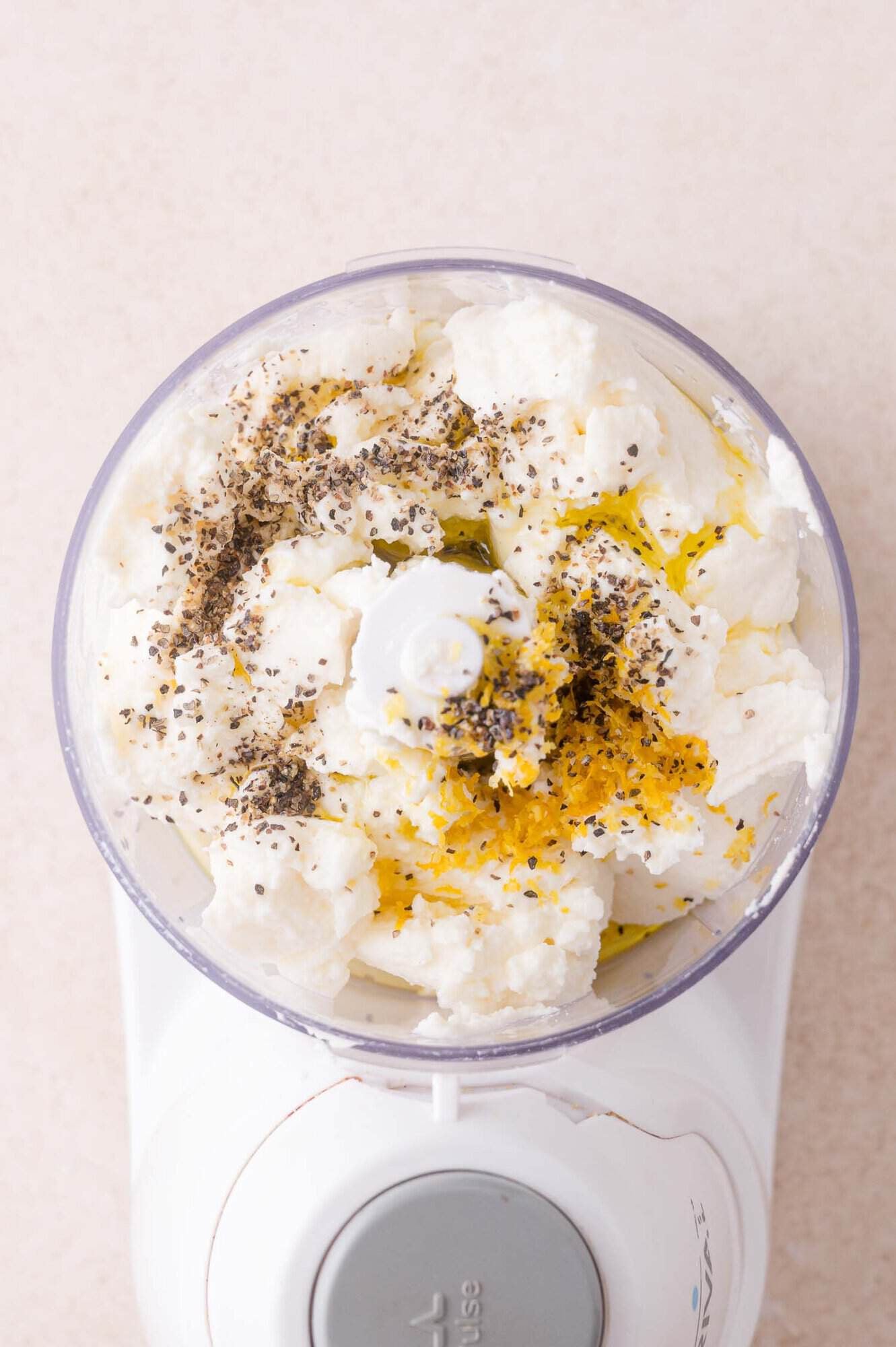 A food processor bowl filled with ricotta cheese, olive oil, lemon zest, and cracked black pepper, ready to be blended.
