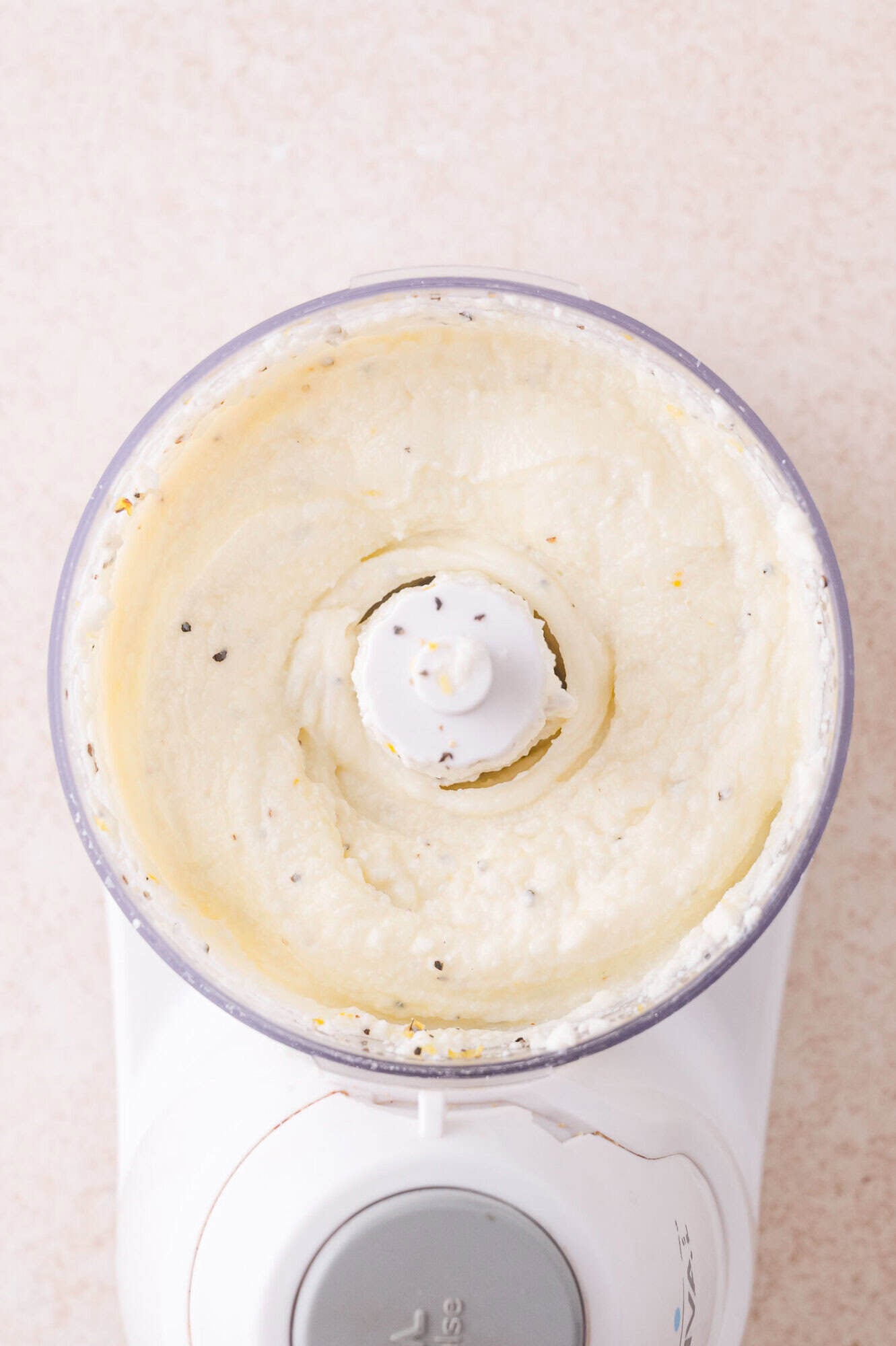 Overhead view of a food processor containing a creamy, blended mixture with visible flecks of seasoning.