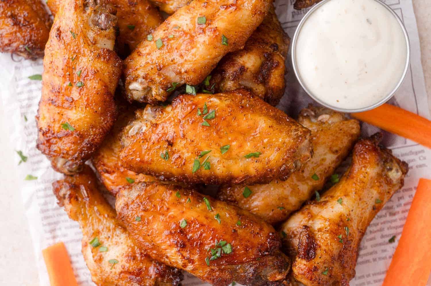 A pile of air fryer chicken wings garnished with herbs, served on newspaper with carrot sticks and a small cup of creamy dipping sauce.