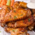 A close-up of a pile of crispy baked chicken wings garnished with herbs, served with celery, carrot sticks, and a cup of ranch dipping sauce.