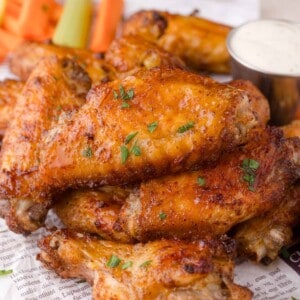 A close-up of a pile of crispy baked chicken wings garnished with herbs, served with celery, carrot sticks, and a cup of ranch dipping sauce.