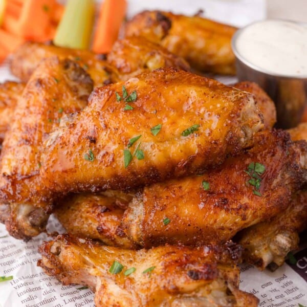 A close-up of a pile of crispy baked chicken wings garnished with herbs, served with celery, carrot sticks, and a cup of ranch dipping sauce.
