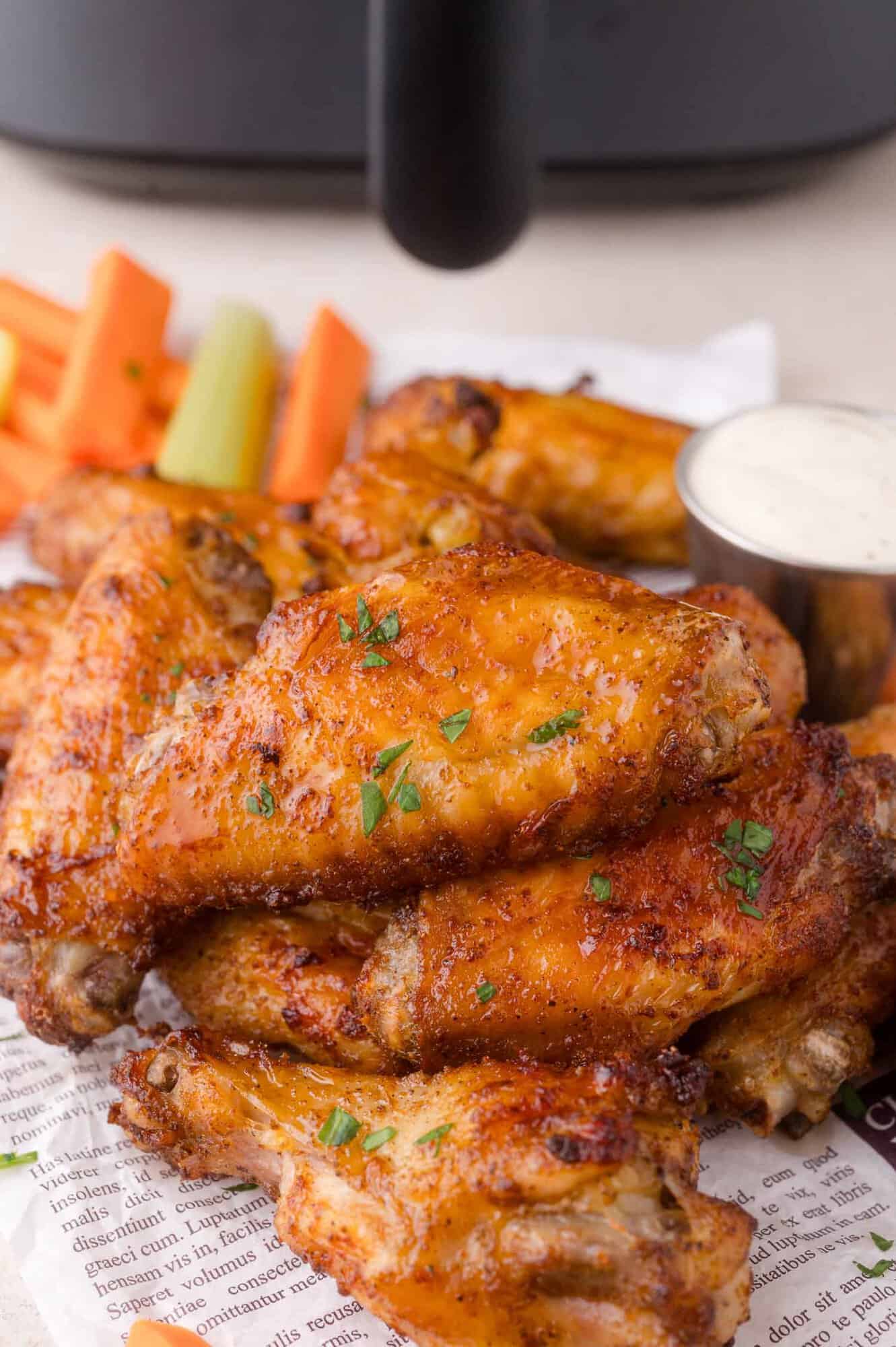 A pile of seasoned chicken wings on newspaper, garnished with herbs, served with ranch dip and celery and carrot sticks in the background.