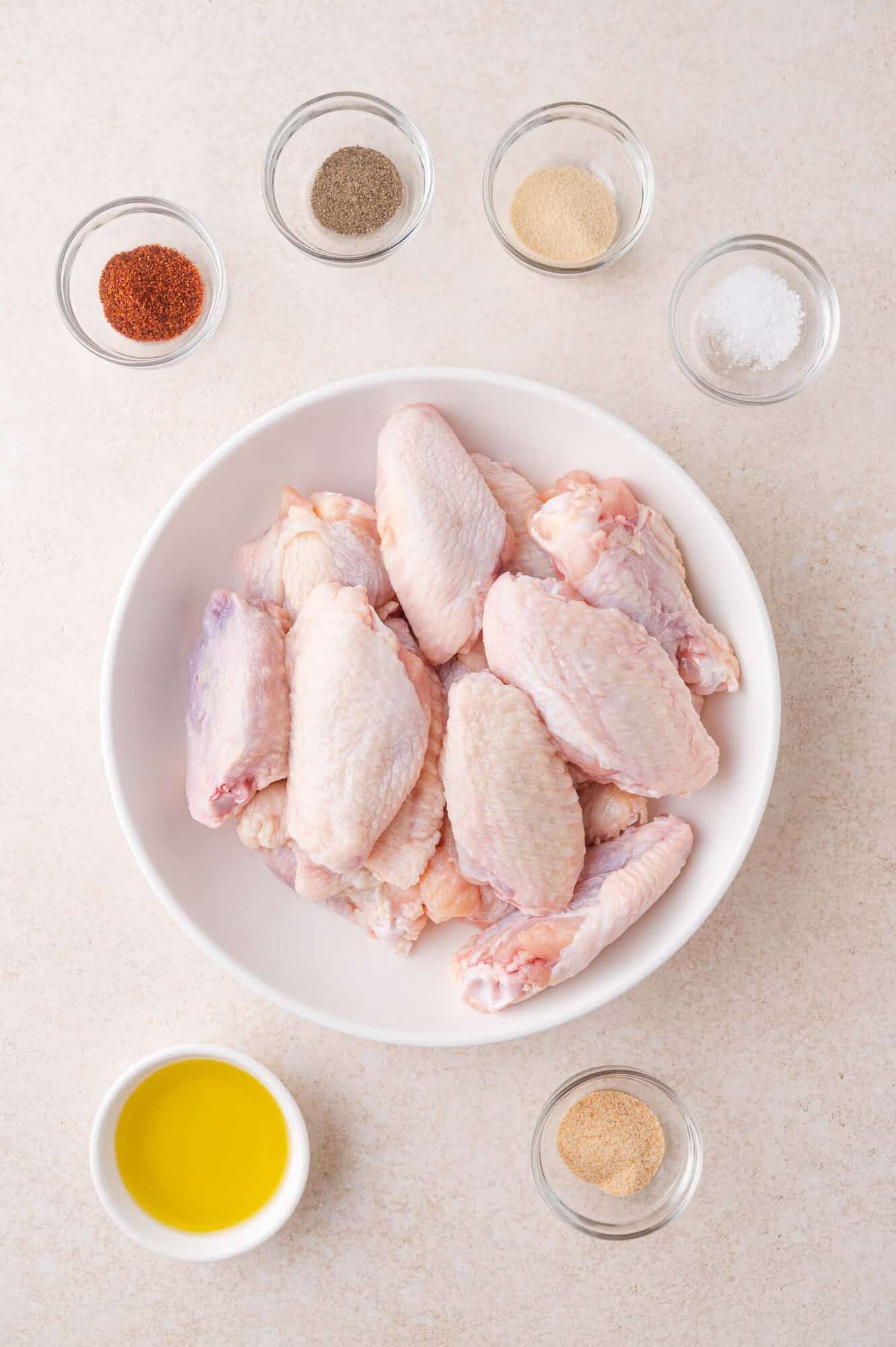 Raw chicken wings in a white bowl surrounded by small bowls of spices, salt, oil, and seasoning on a light-colored surface.