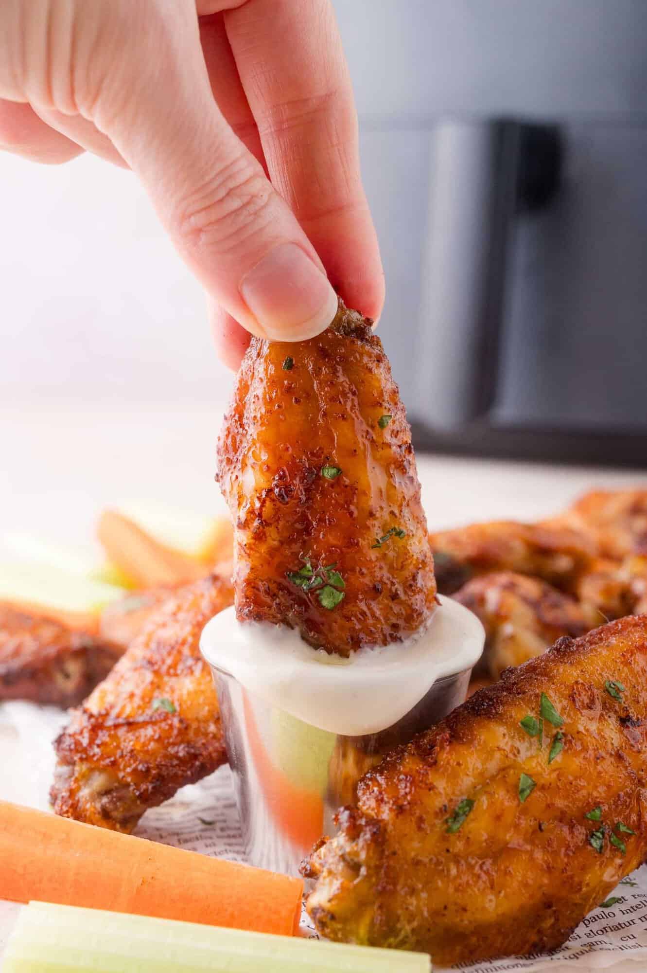 A hand dips a seasoned chicken wing into a cup of ranch sauce, with more wings, carrot sticks, and celery on a tray in the background.