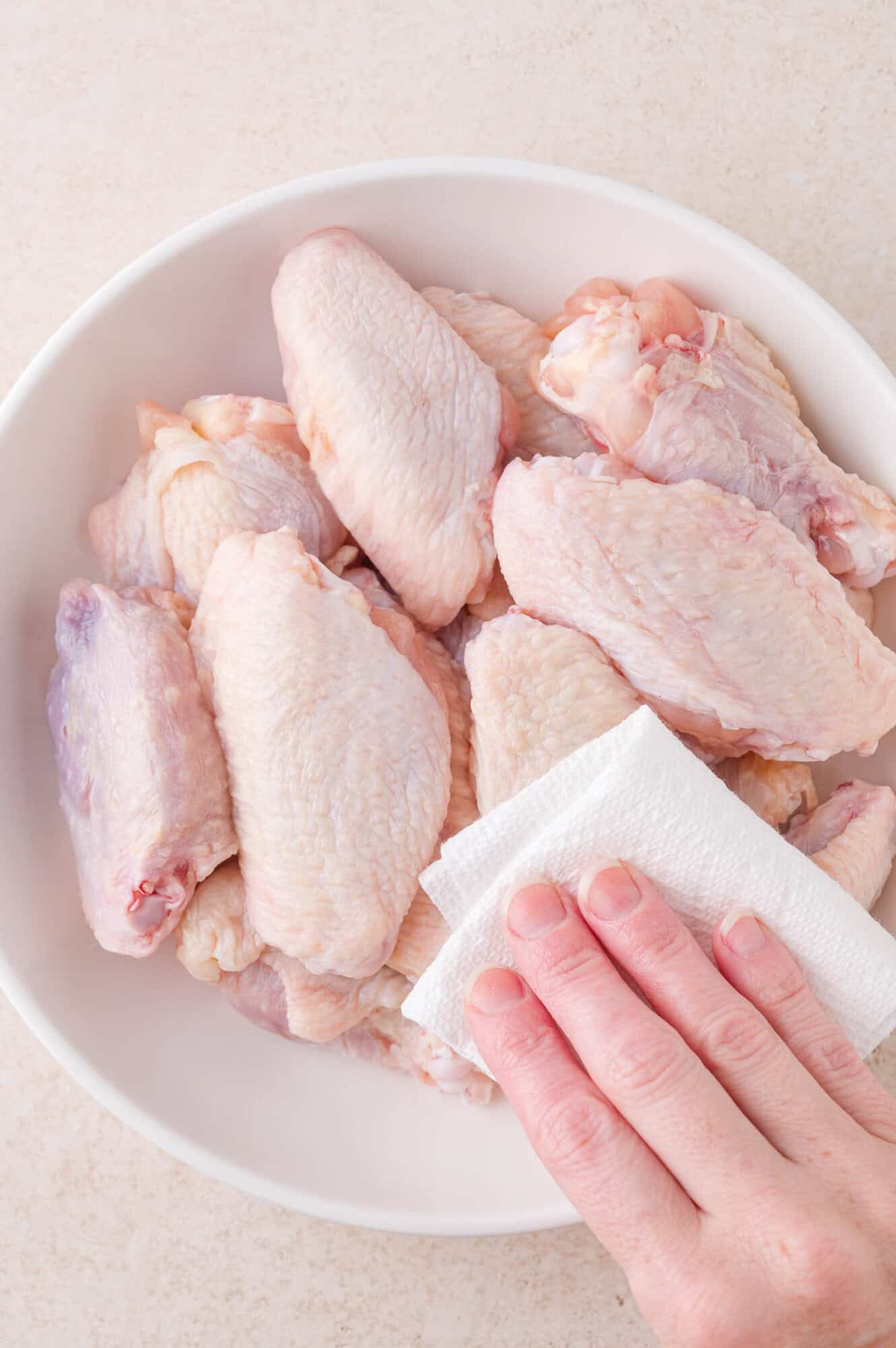 A hand uses a paper towel to pat dry raw chicken wings in a white bowl.