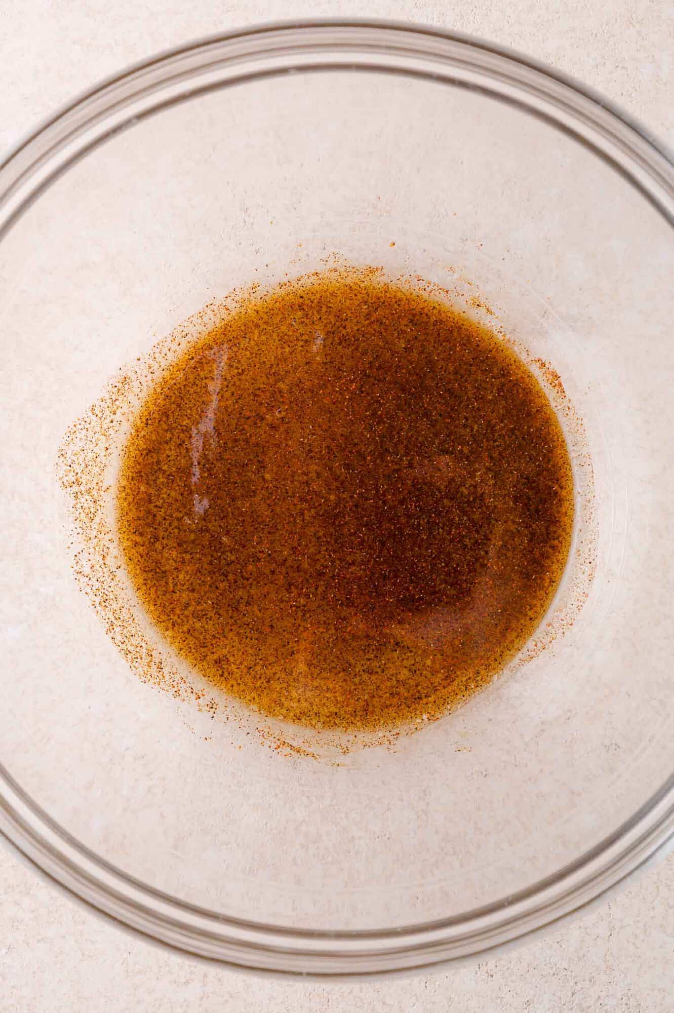 A clear glass bowl containing a mixture of oil and spices sits on a light-colored surface.
