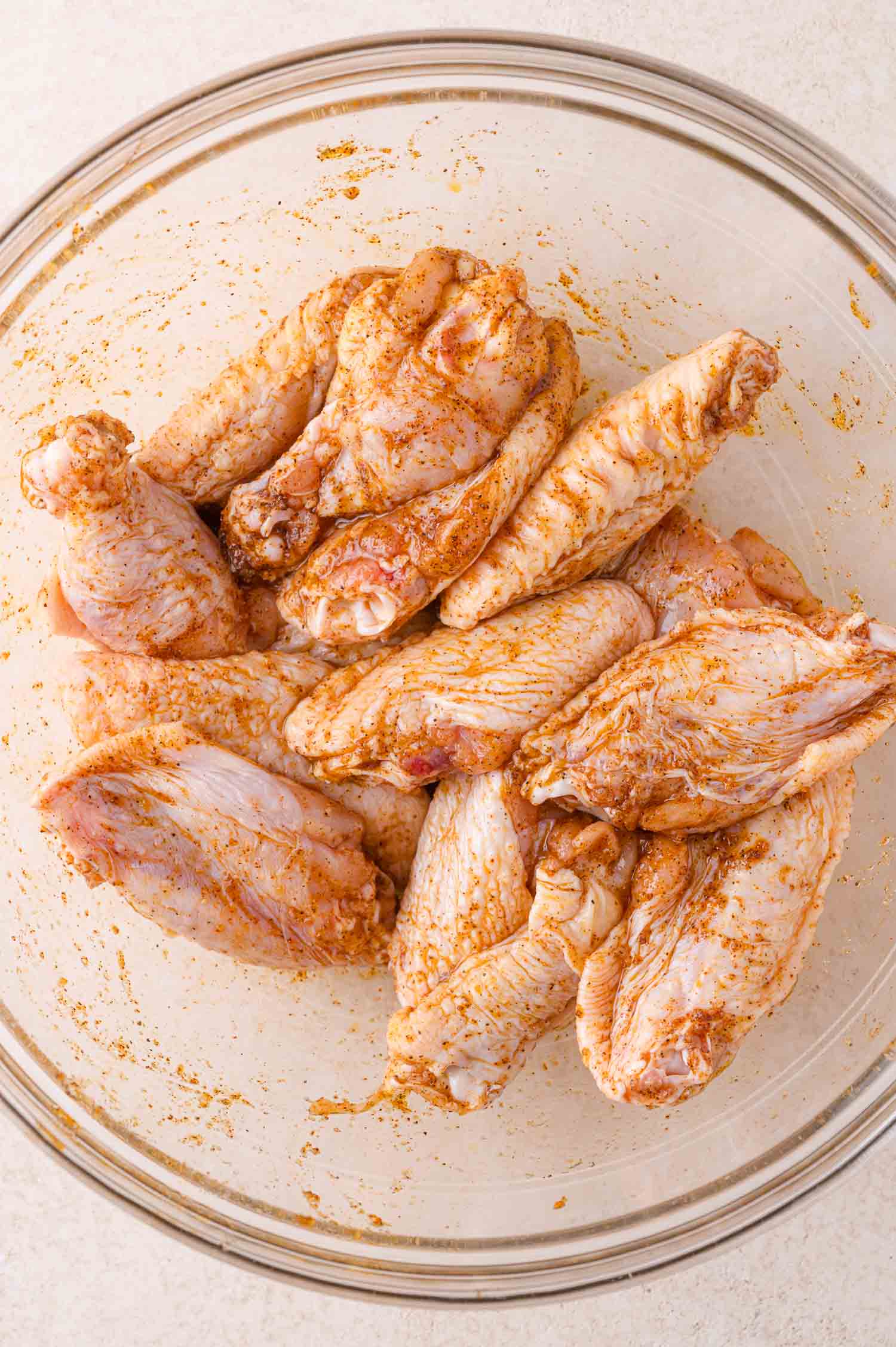 Raw chicken wings coated with seasoning are arranged in a large clear glass bowl on a light-colored surface.