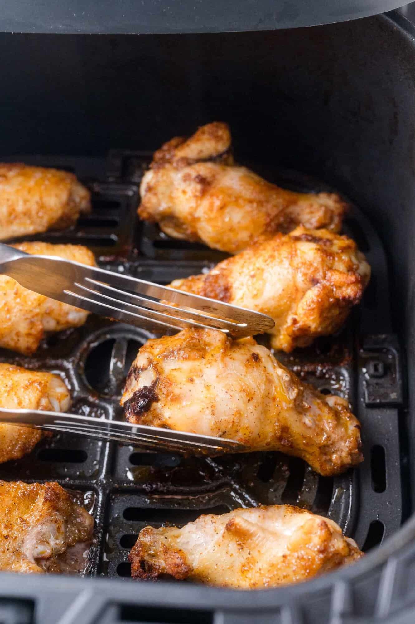 Several cooked chicken wings in an air fryer basket, with a fork and tongs lifting one wing.