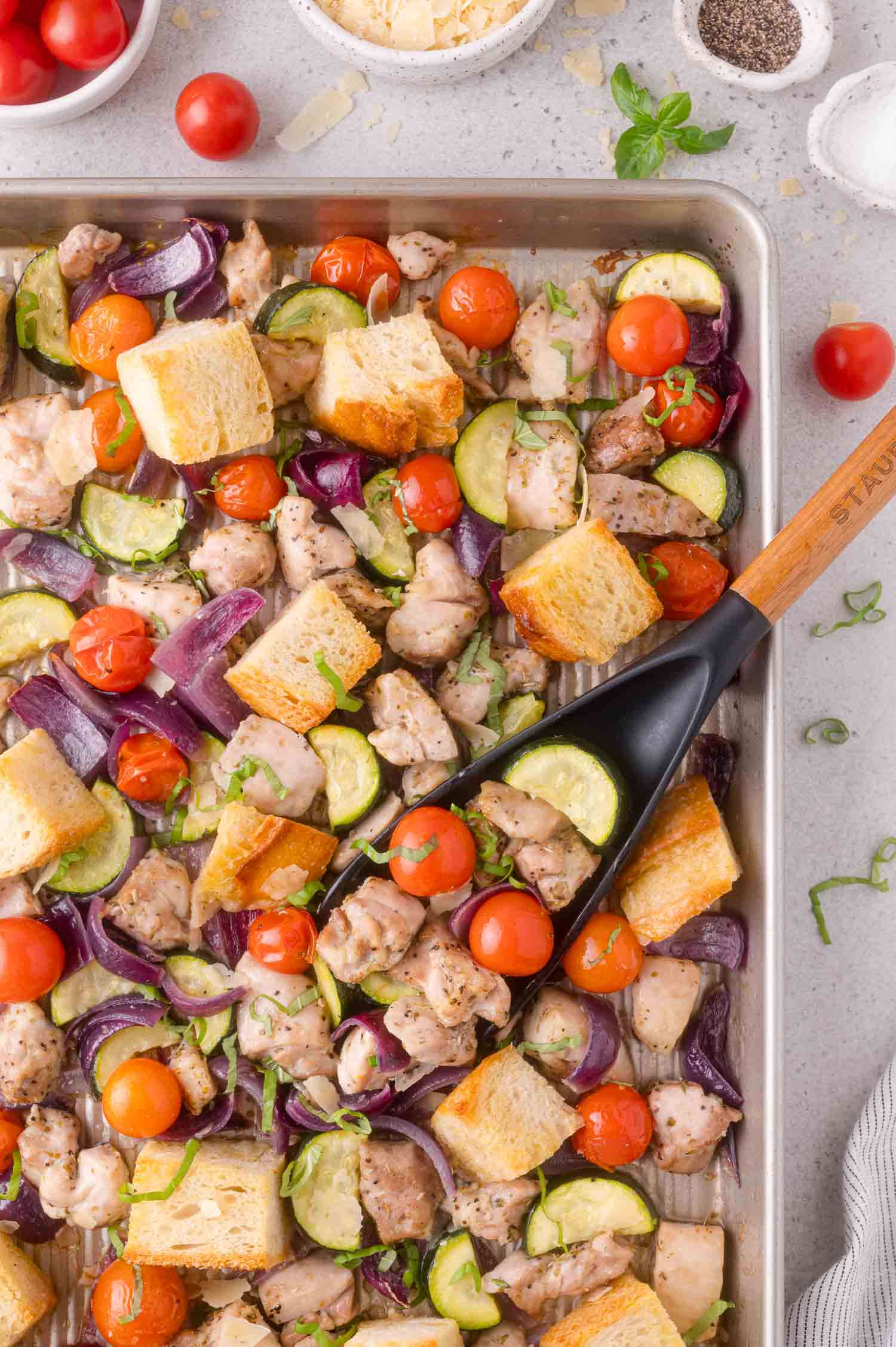 Sheet pan with roasted chicken, cherry tomatoes, zucchini, red onion, and bread cubes, with a spatula lifting a portion.