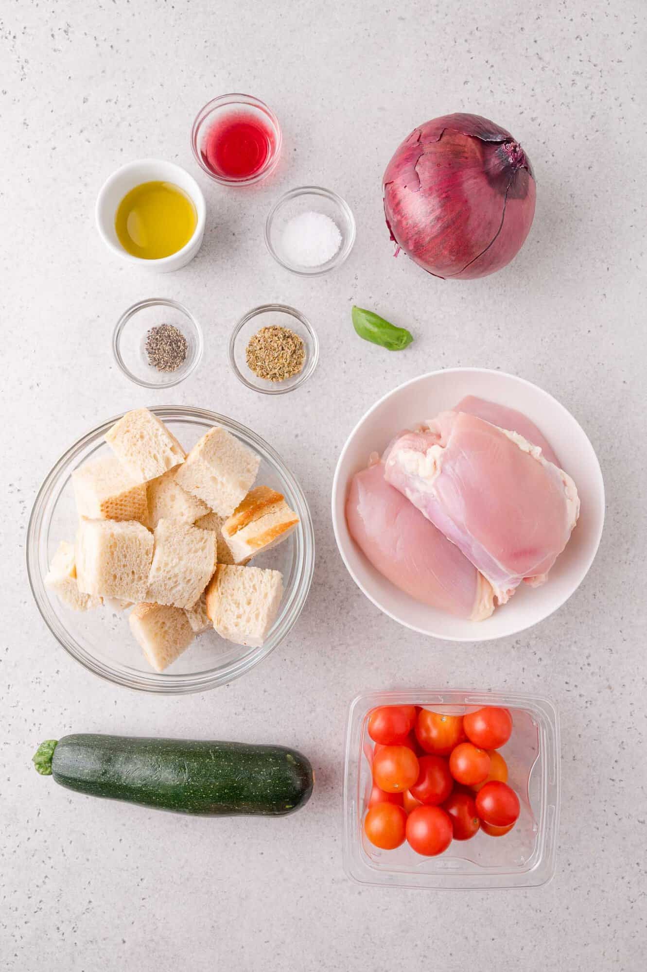 Ingredients on a countertop, including chicken thighs, cubed bread, cherry tomatoes, zucchini, red onion, basil, olive oil, vinegar, salt, pepper, and dried herbs.