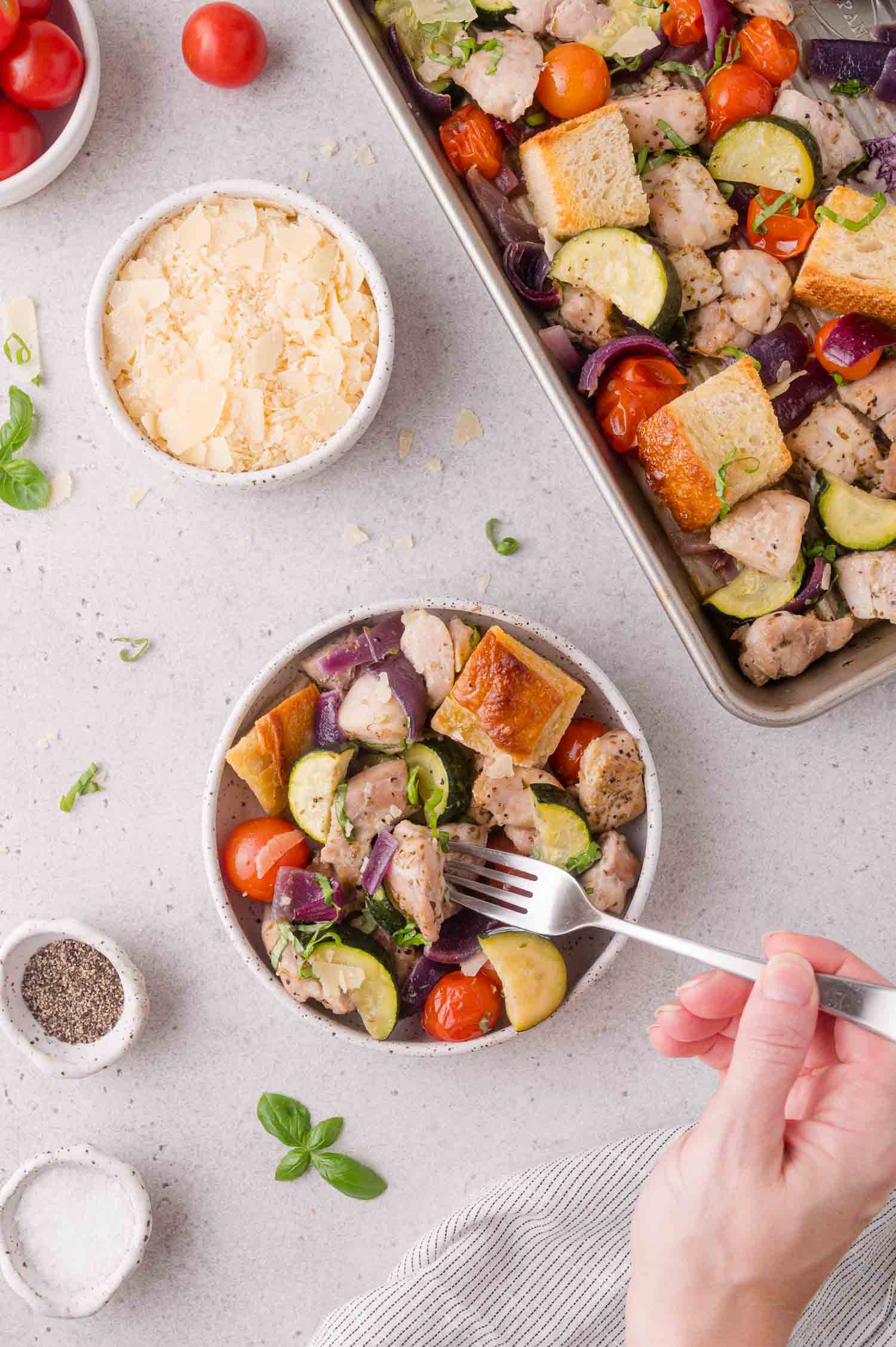 A hand holds a fork over a bowl of salad with chicken, bread cubes, zucchini, tomatoes, and red onions. Nearby are bowls of shredded cheese, salt, pepper, and a tray of the same salad.