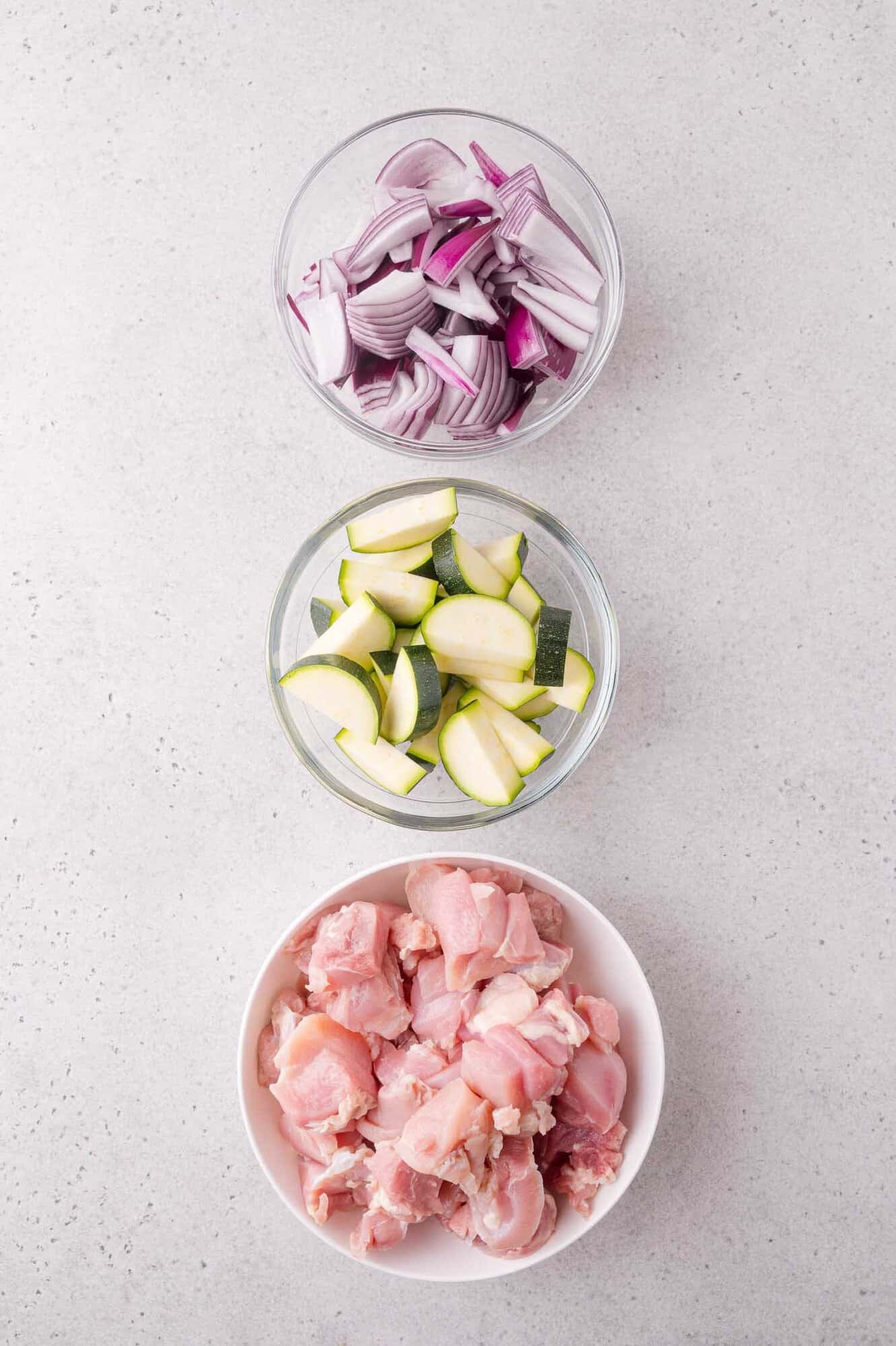 Three bowls containing chopped red onions, sliced zucchini, and raw diced chicken arranged vertically on a light gray surface.