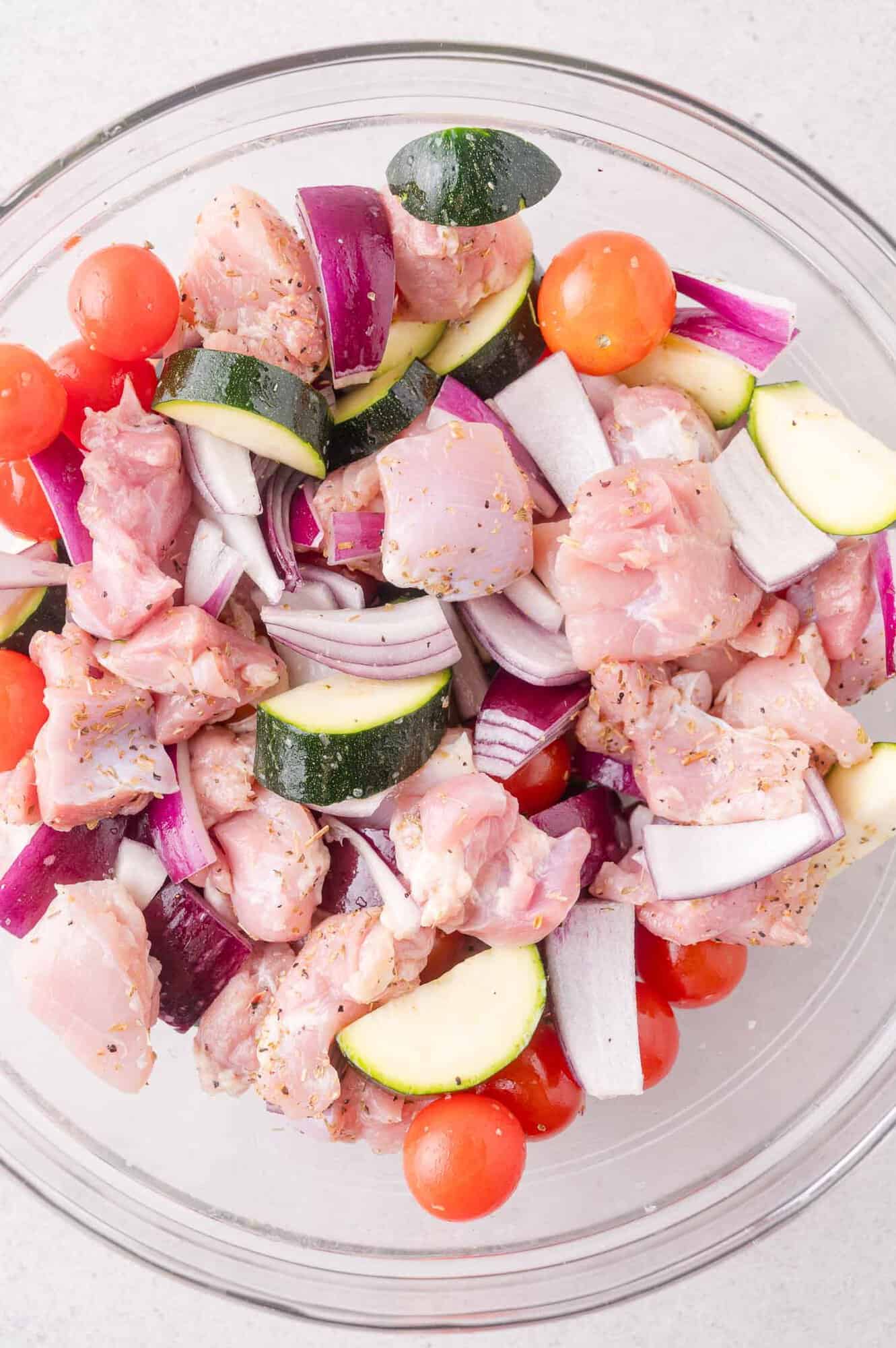 A glass bowl filled with raw chicken pieces, cherry tomatoes, zucchini slices, and red onion chunks, all seasoned and ready for cooking.
