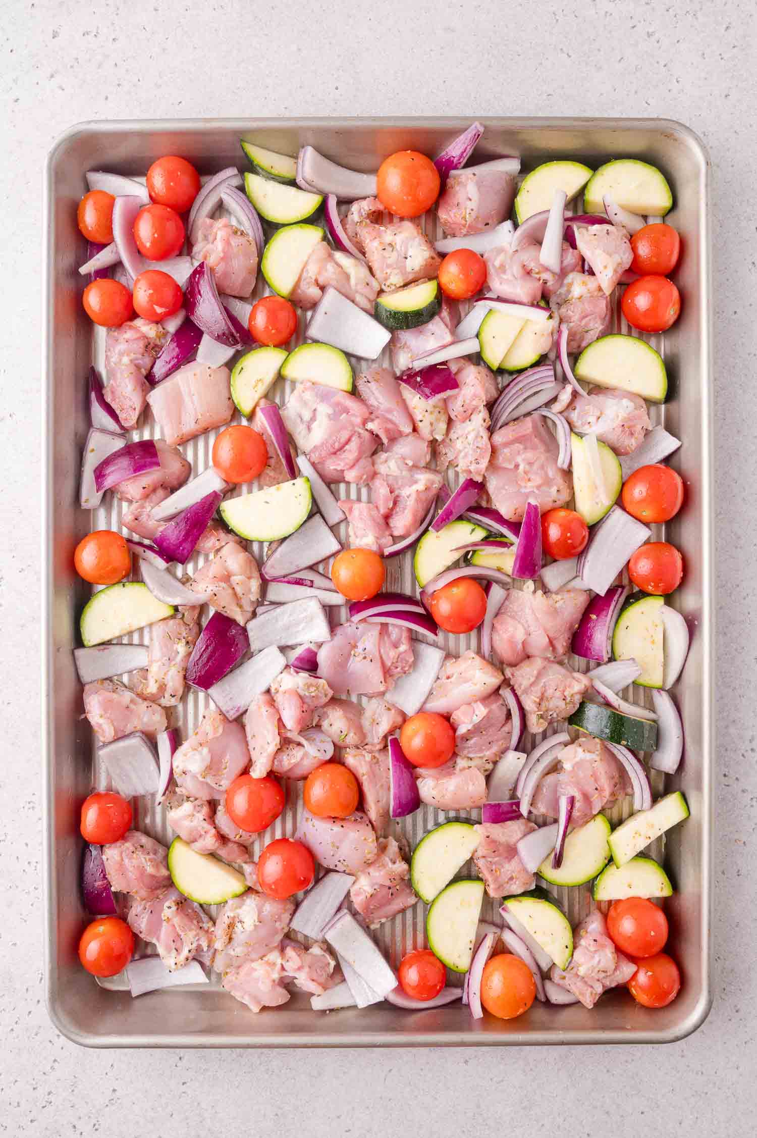 Raw chicken pieces, cherry tomatoes, red onion slices, and zucchini chunks arranged on a metal baking sheet, ready for roasting.