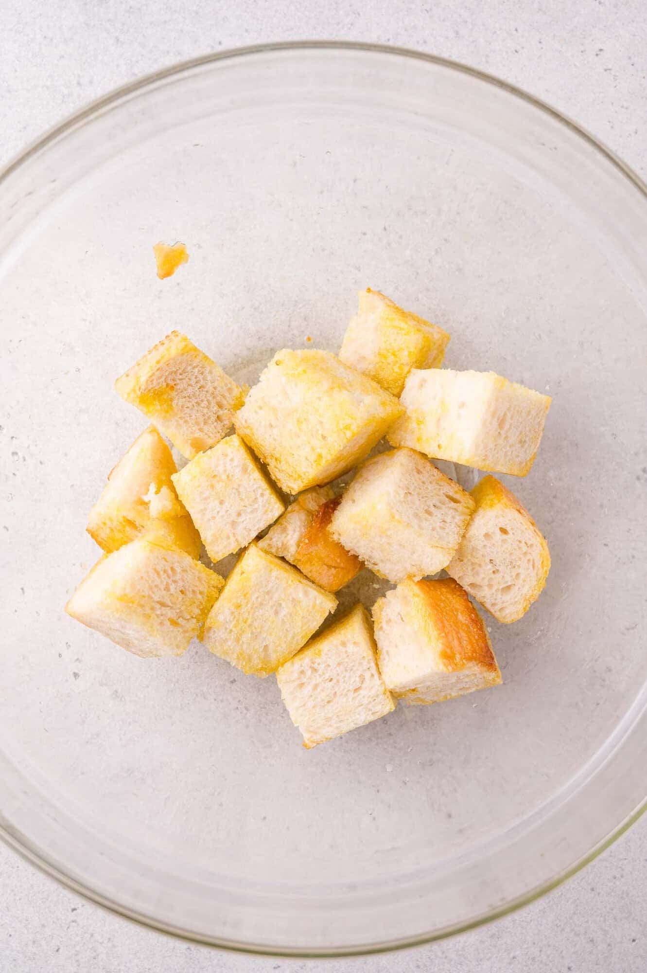 Cubed pieces of bread coated with oil or butter in a clear glass mixing bowl on a light-colored surface.