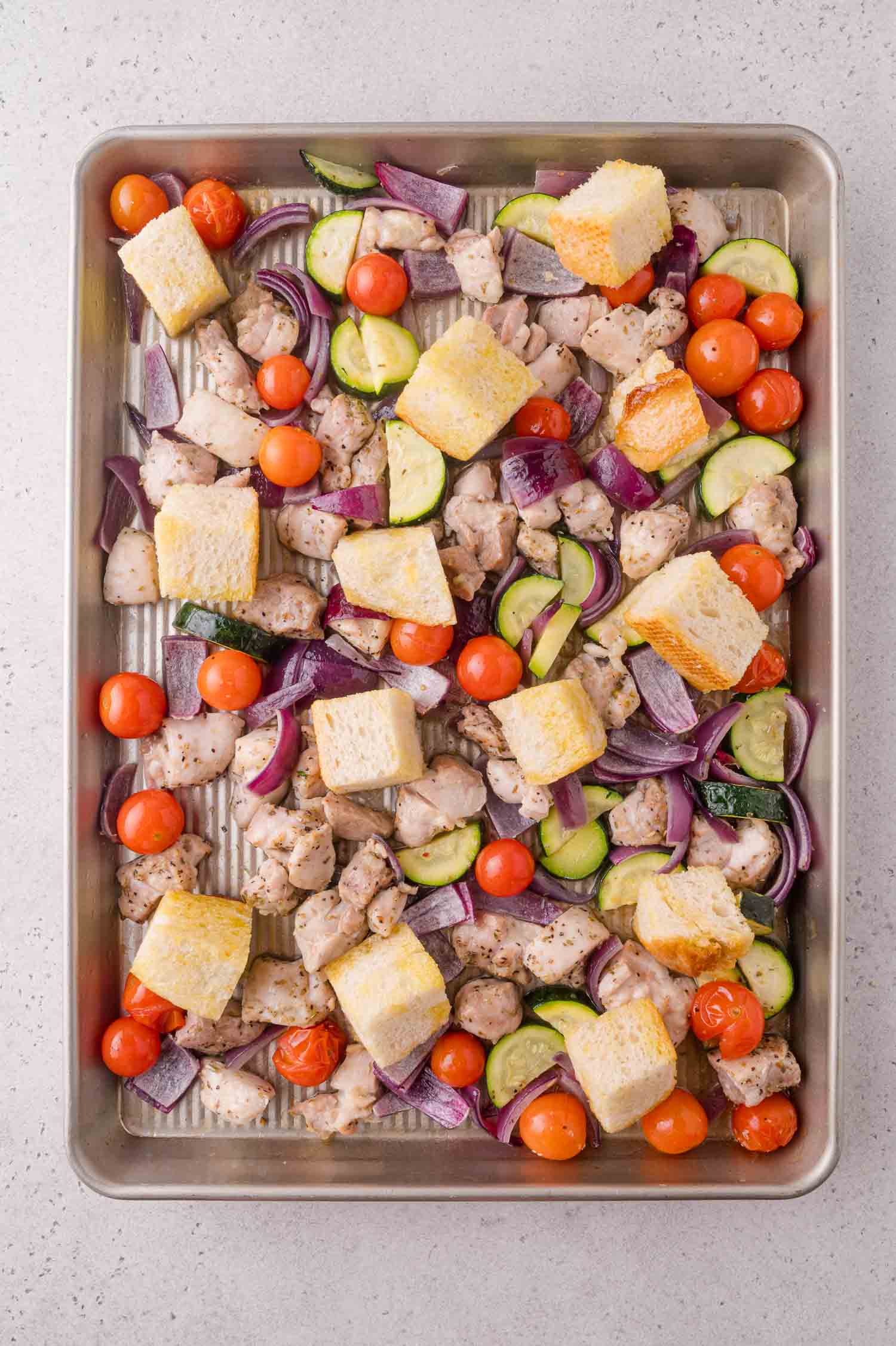 Sheet pan with diced chicken, cherry tomatoes, zucchini slices, red onion pieces, and cubes of bread, all seasoned and ready to be roasted.