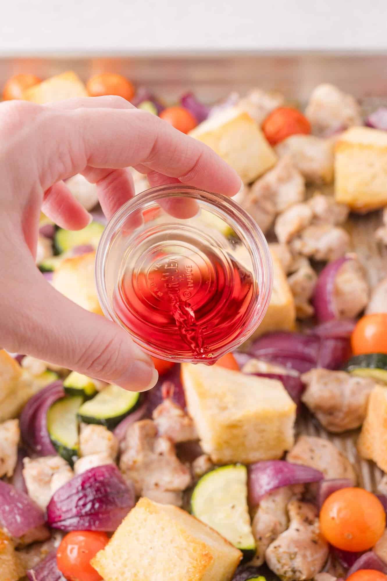 A hand pours red wine vinegar from a small glass bowl over a sheet pan filled with chopped vegetables, bread cubes, and pieces of chicken.