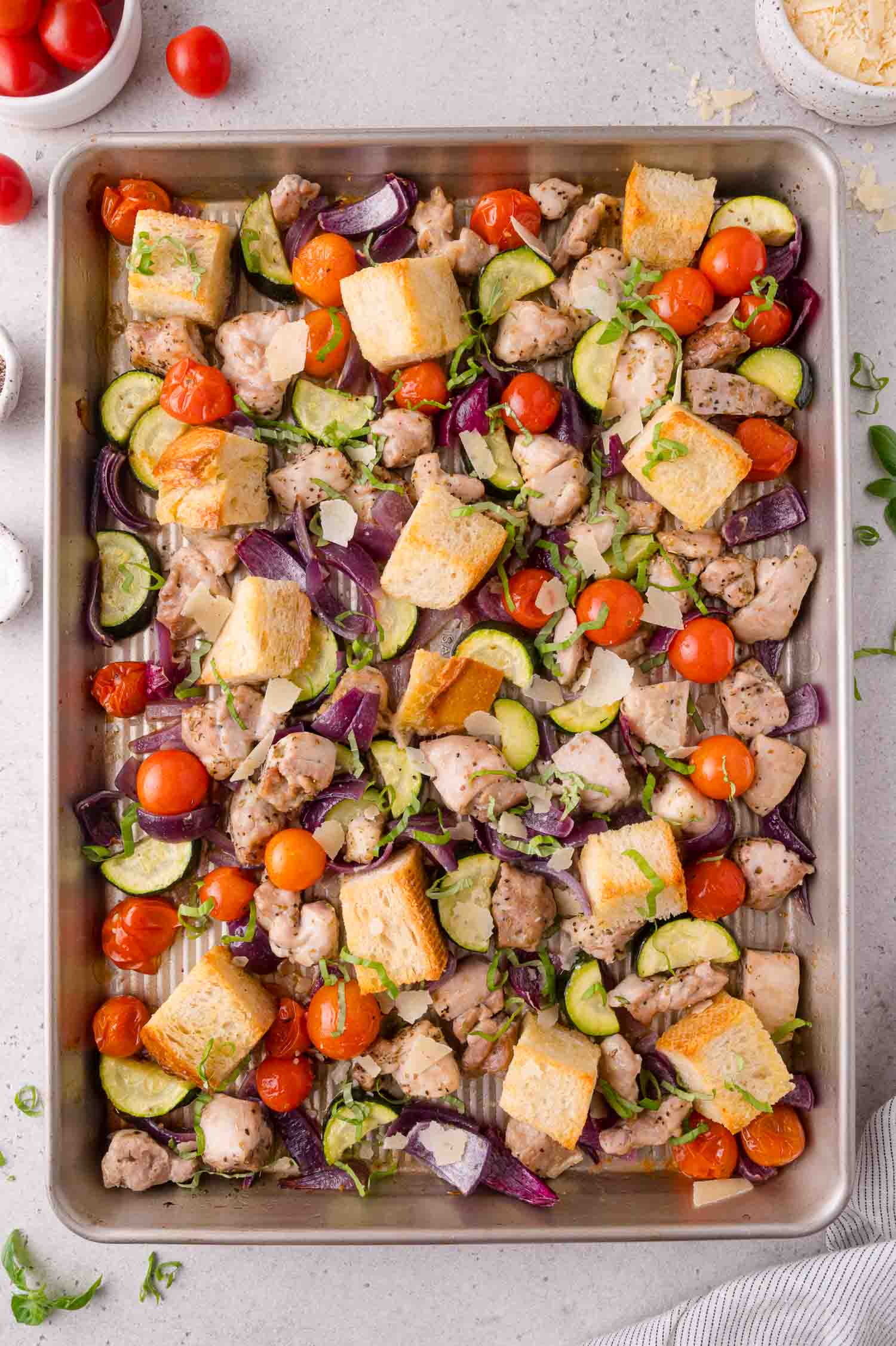 A sheet pan filled with roasted chicken pieces, cherry tomatoes, zucchini, red onion, and cubes of bread, garnished with herbs.