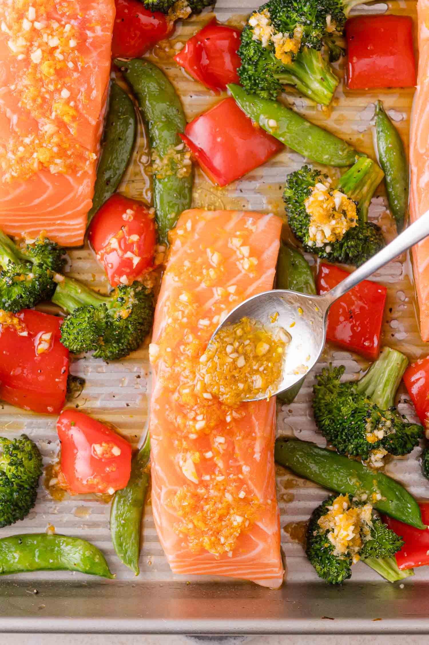 Uncooked salmon fillets on a baking sheet with broccoli, red bell pepper, and snap peas, being drizzled with a garlic and ginger sauce.