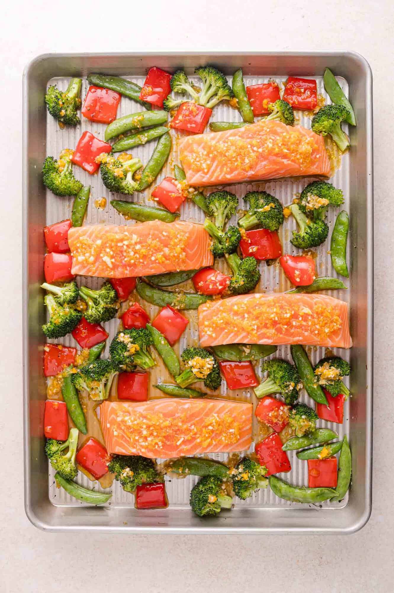 Raw salmon fillets, broccoli, snap peas, and red bell peppers arranged on a metal baking tray, ready for cooking.