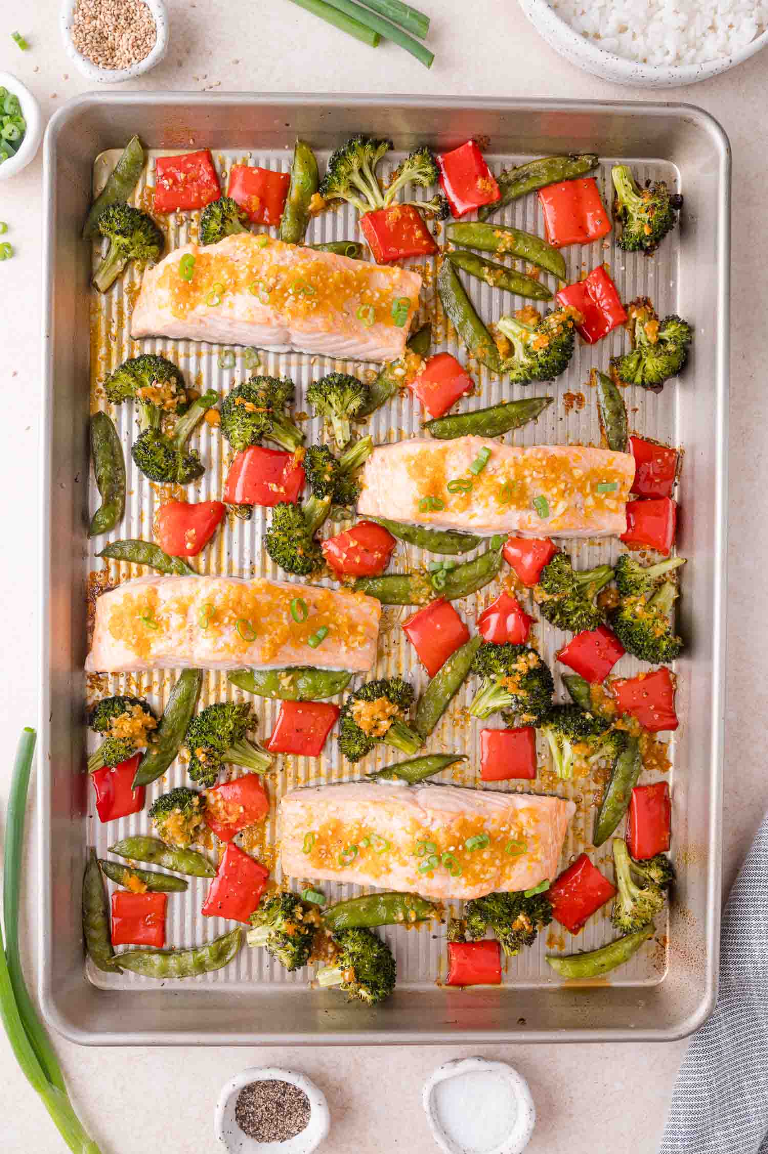 Baked salmon fillets on a sheet pan with broccoli, red bell peppers, and snap peas, garnished with sesame seeds and green onions.