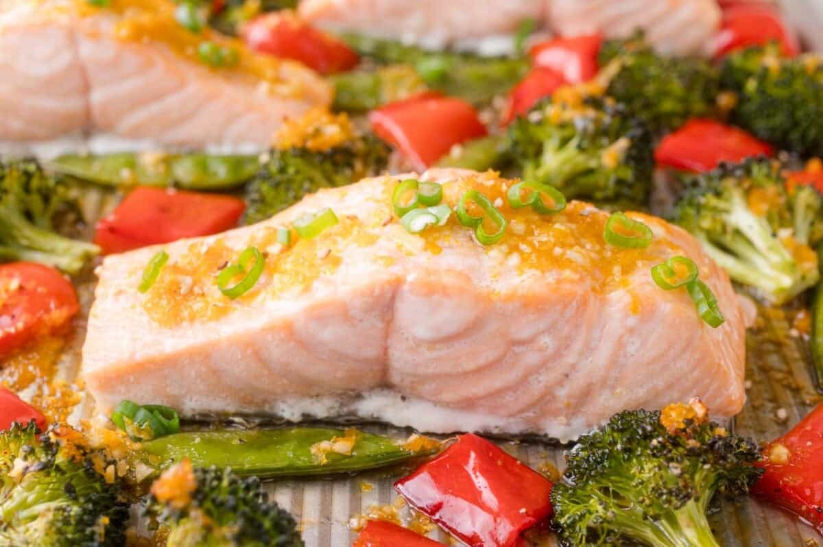 Baked salmon fillet topped with chopped green onions, surrounded by broccoli florets, red bell peppers, and snap peas on a sheet pan.