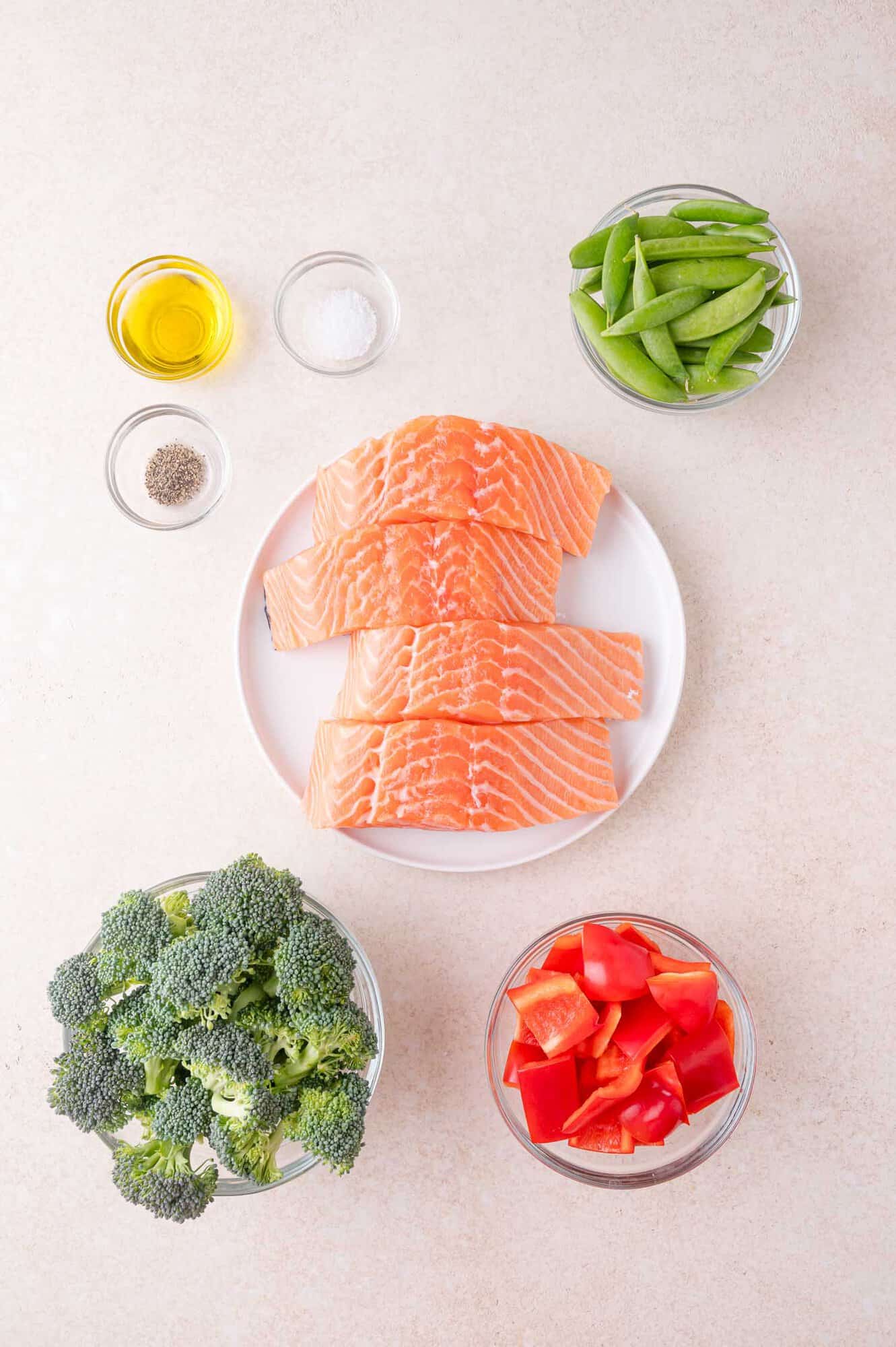 A plate of raw salmon fillets on a white surface, surrounded by bowls of broccoli, red bell pepper, snap peas, olive oil, salt, and pepper.